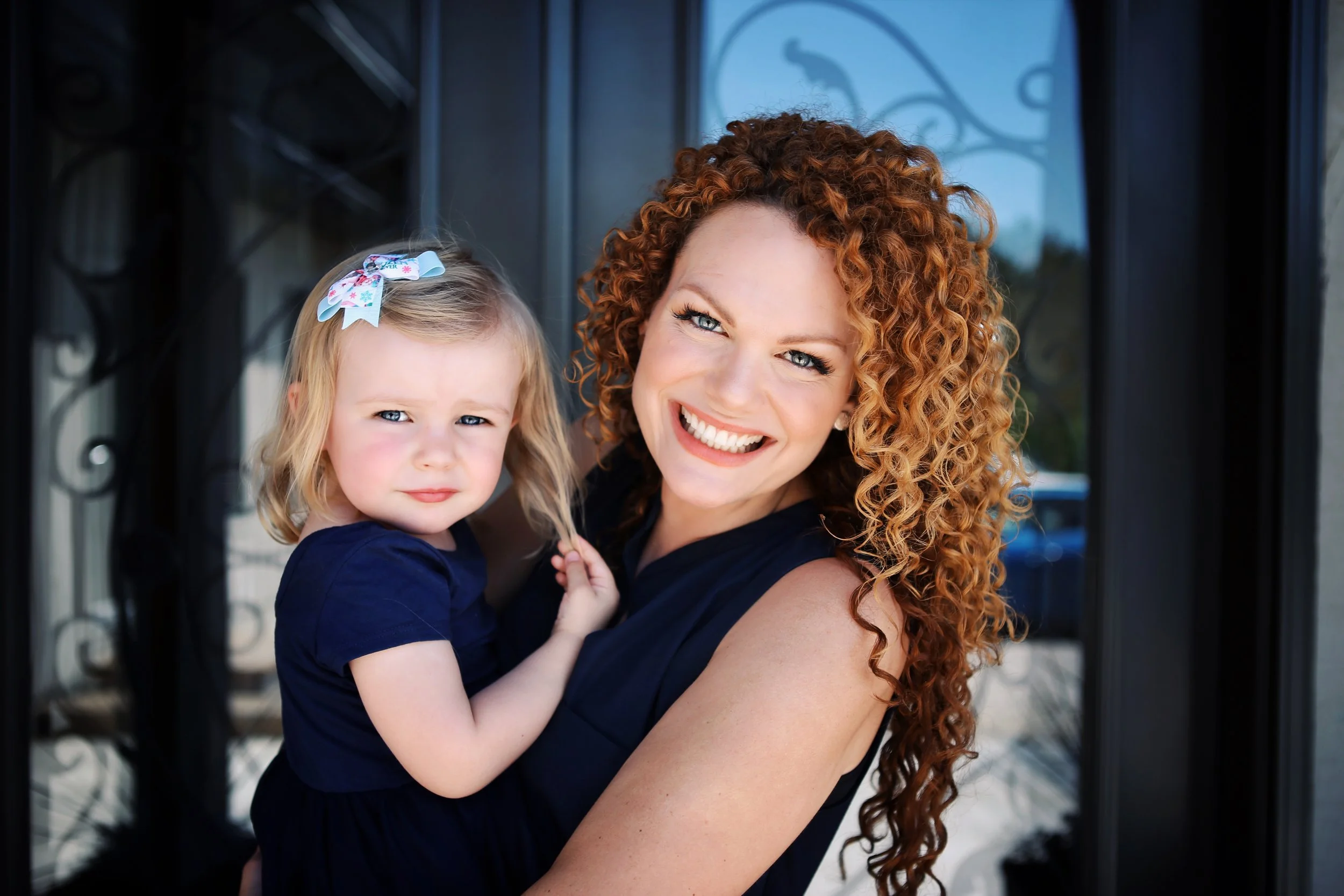 A woman with curly red hair holding a young girl with blonde hair and a bow, both smiling outdoors in front of a decorative black door.