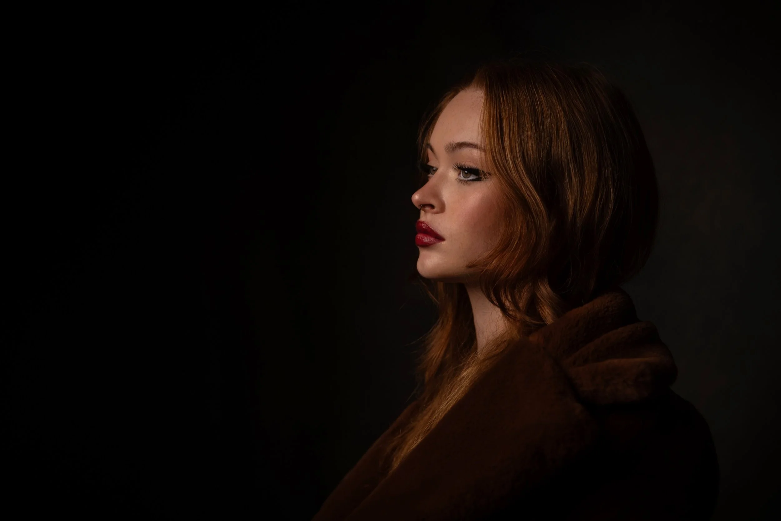 Side profile of a woman with reddish-brown hair, wearing a brown coat, against a dark background.