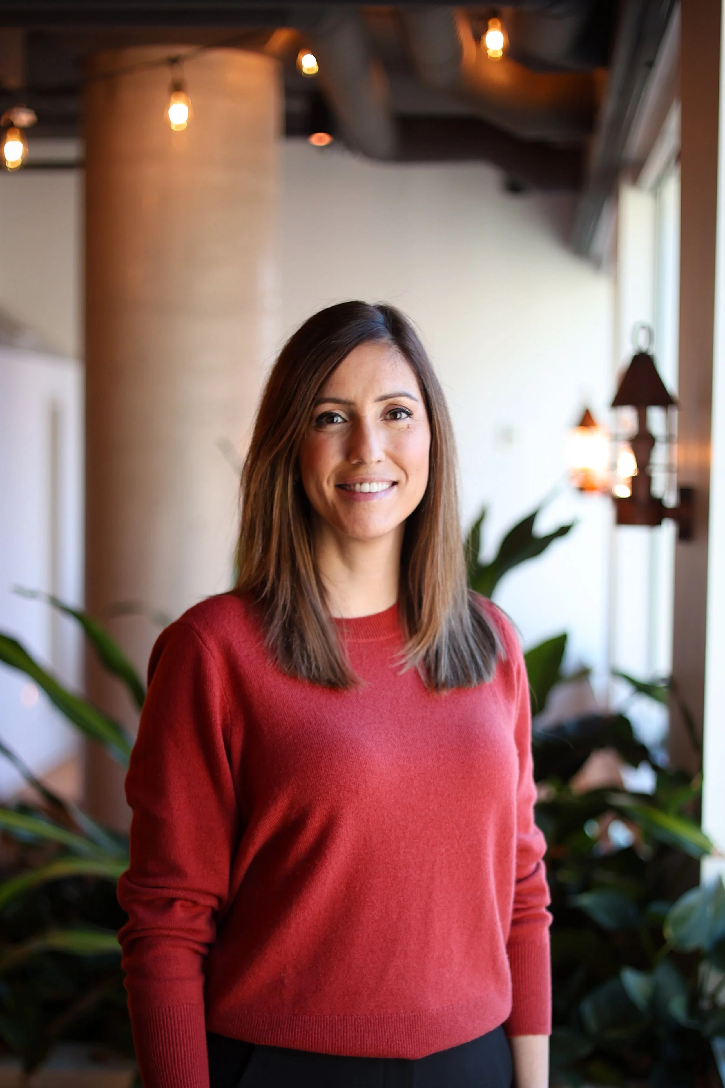 A woman with shoulder-length brown hair wearing a red sweater, standing indoors with natural light from a window on the right, with plants and warm lighting in the background.