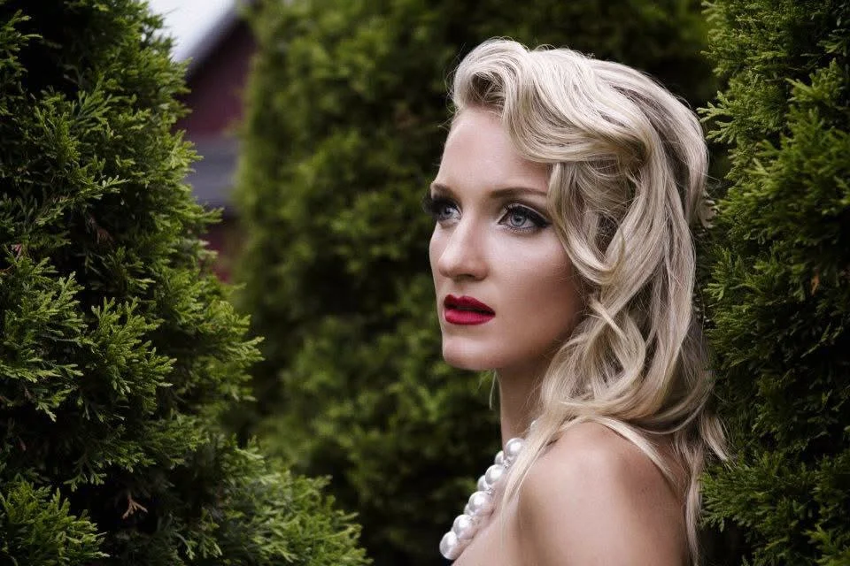 A woman with blonde hair styled in waves and red lipstick gazes thoughtfully while standing between tall green bushes.