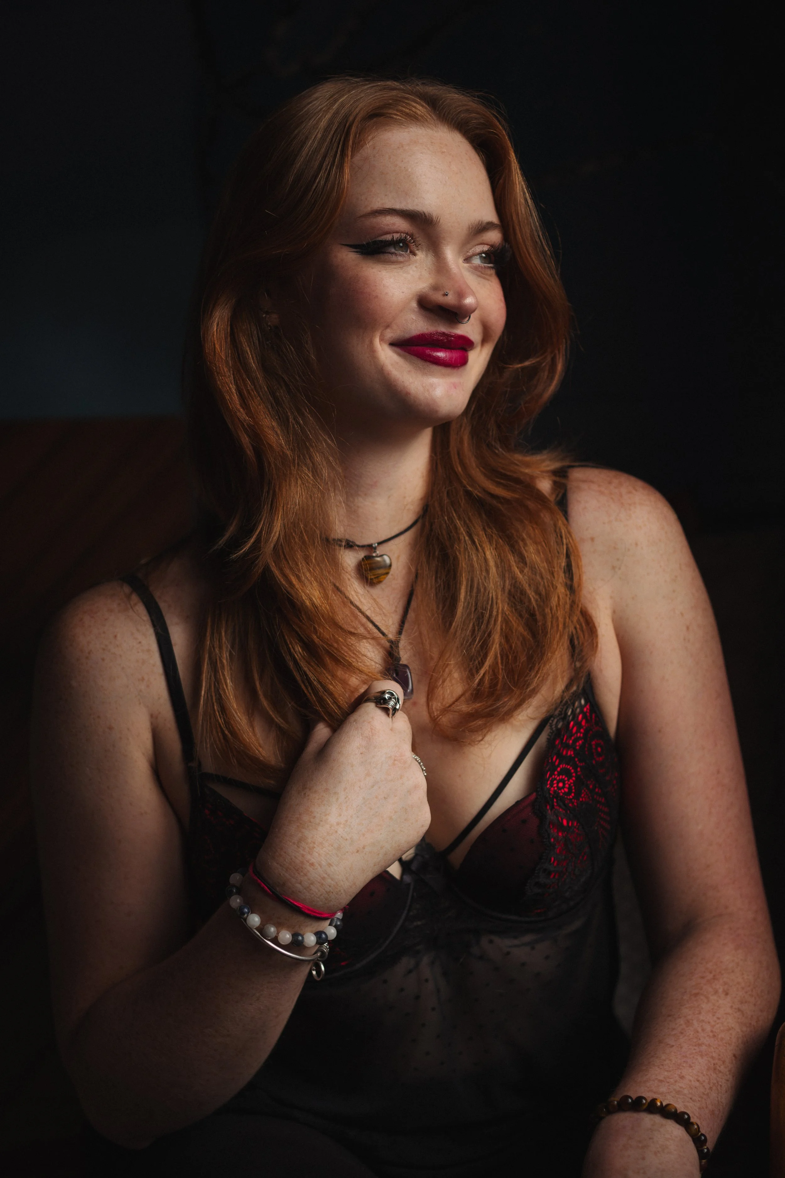 A young woman with red hair, freckles, and makeup is smiling and looking to the side. She is wearing jewelry and a black lace top with red accents.