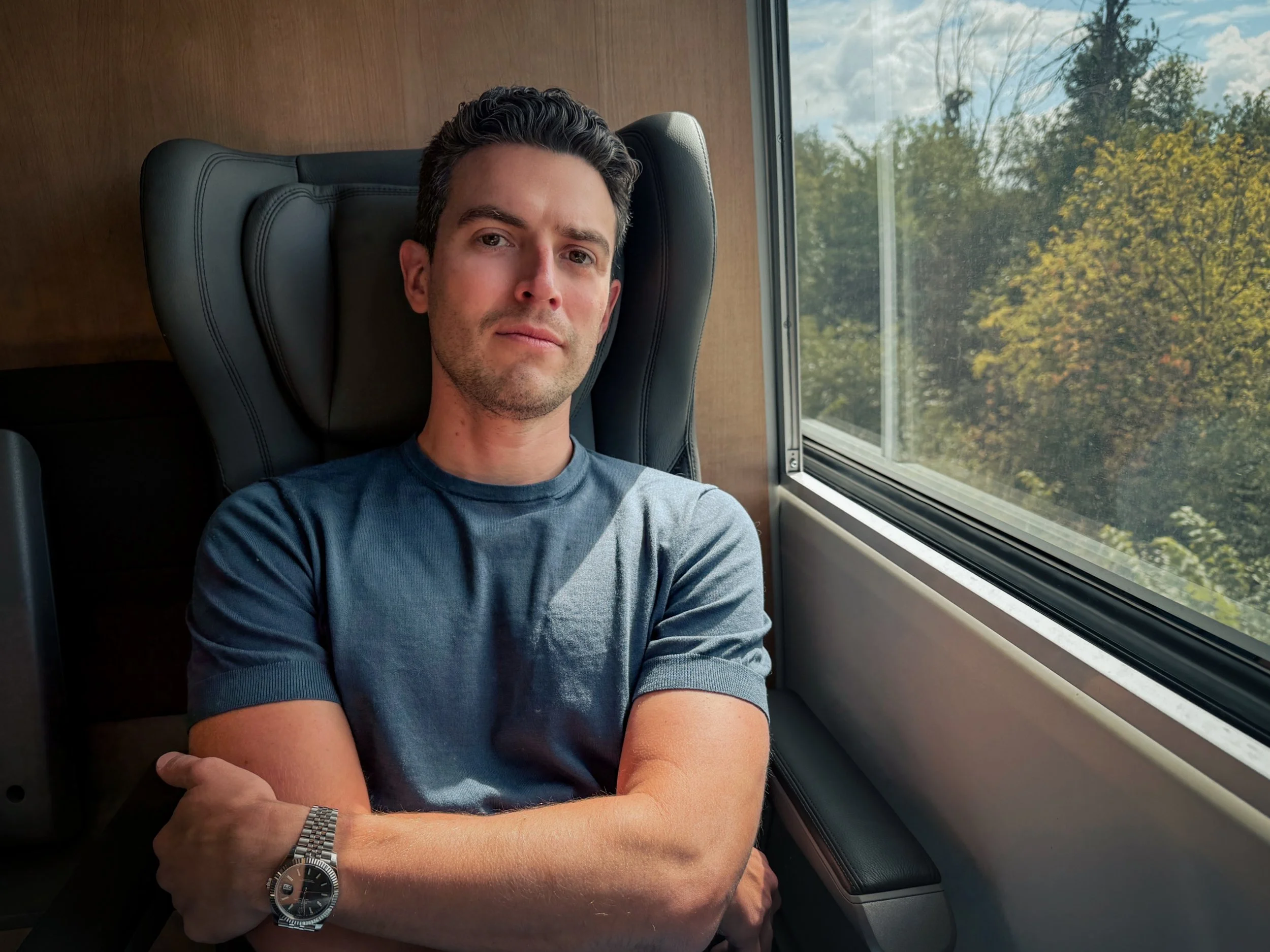 A man sitting on a train or bus, looking at the camera, with arms crossed, wearing a blue t-shirt and a silver watch, next to a window showing trees and a cloudy sky outside.