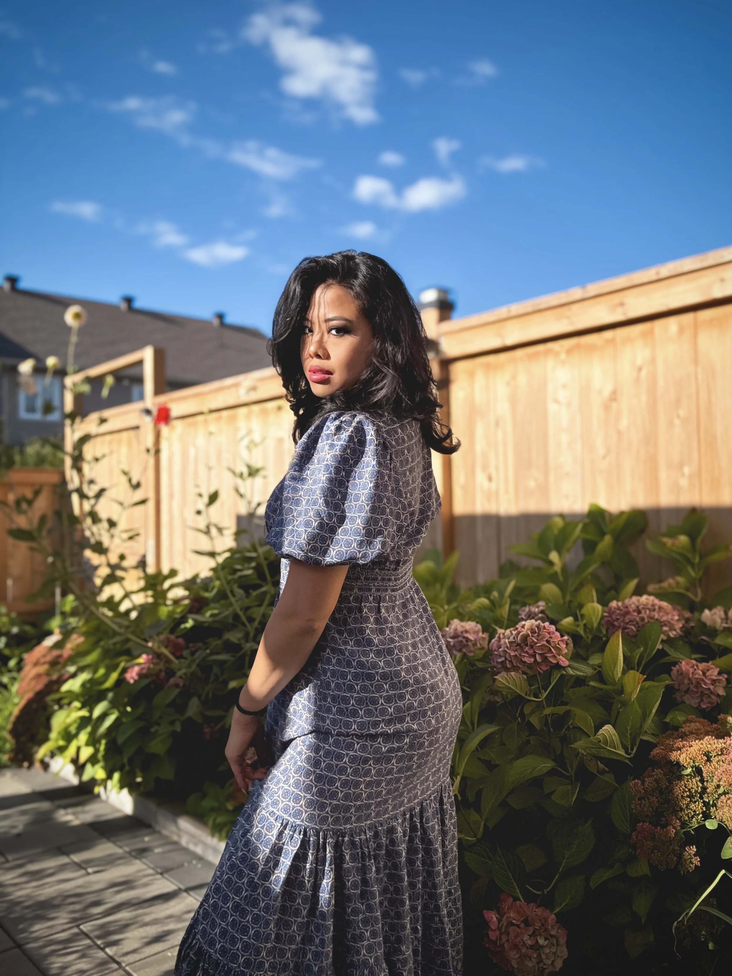 A woman with dark wavy hair and red lipstick stands in a backyard garden, wearing a patterned dress. She looks over her shoulder towards the camera with a serious expression. There are hydrangea bushes with pink flowers behind her, a wooden fence, an