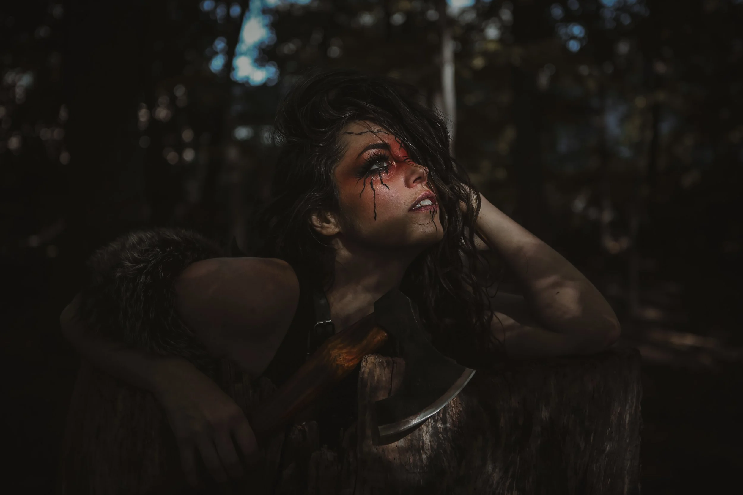 A woman with dark, curly hair and face paint resembling a bleeding eye, leaning on a tree stump with an axe resting on it, in a dark forest setting.