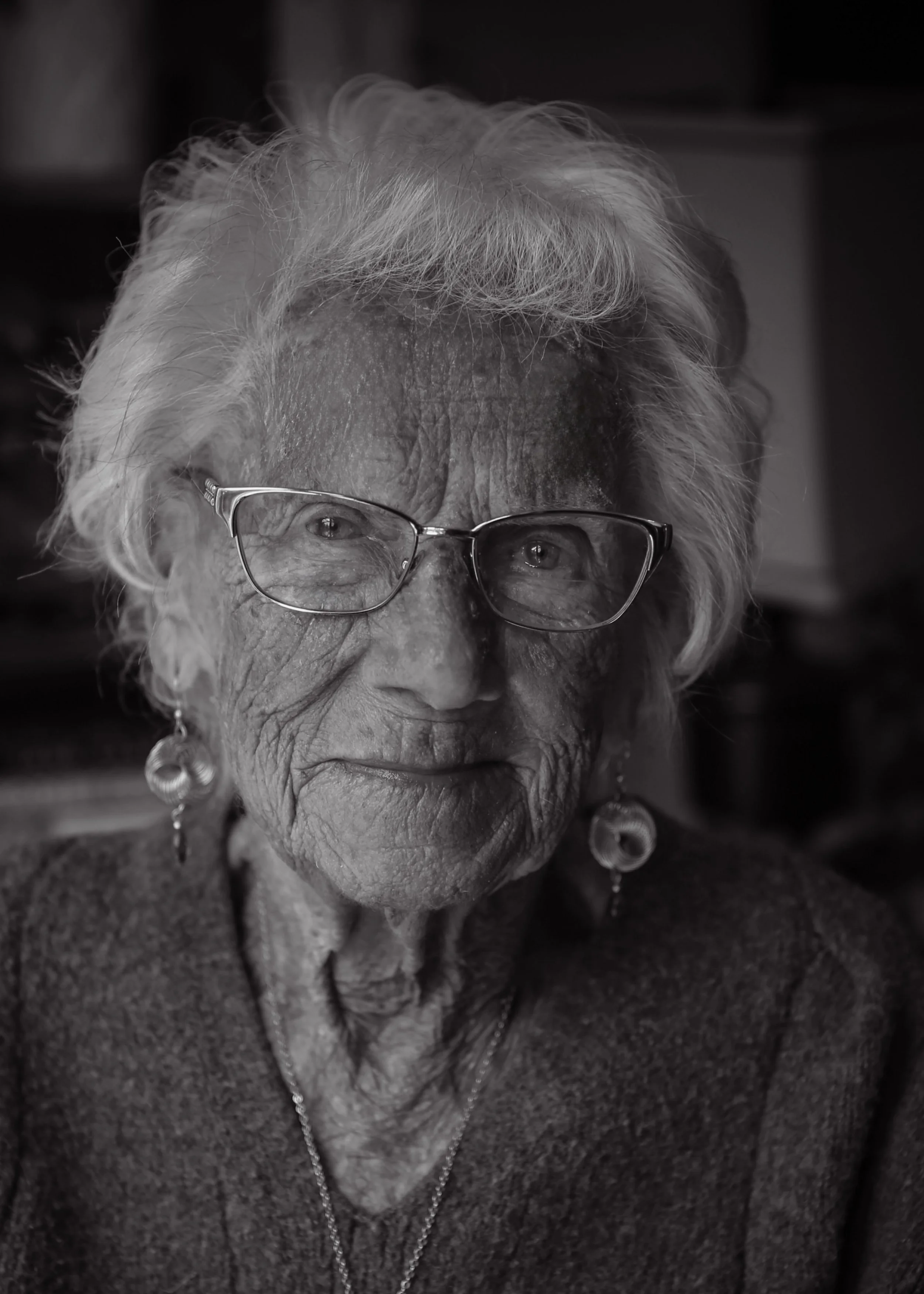 Close-up black and white portrait of elderly woman with glasses, earrings, and necklace.