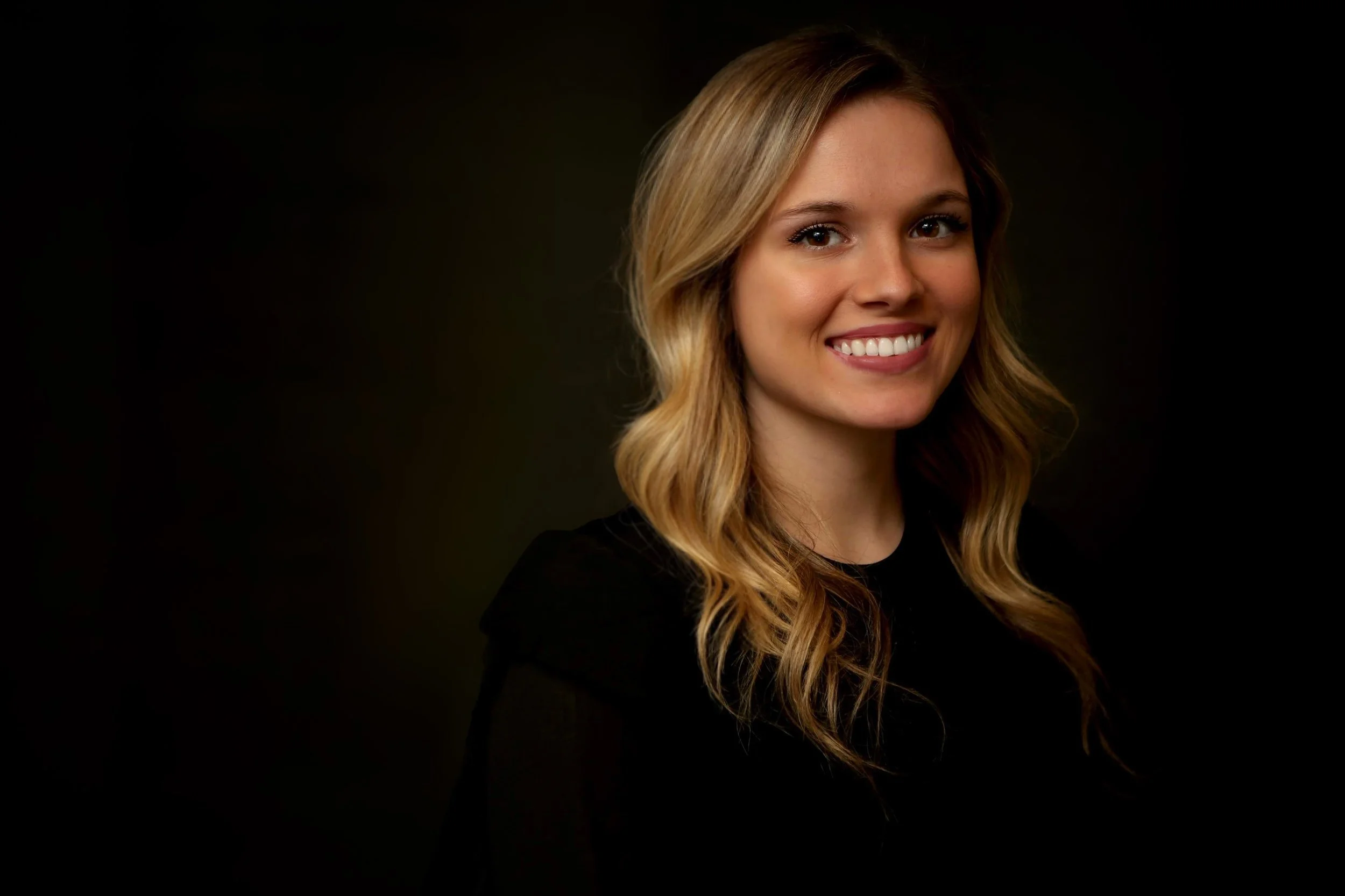 Portrait of a young woman with long, wavy blonde hair smiling against a dark background.