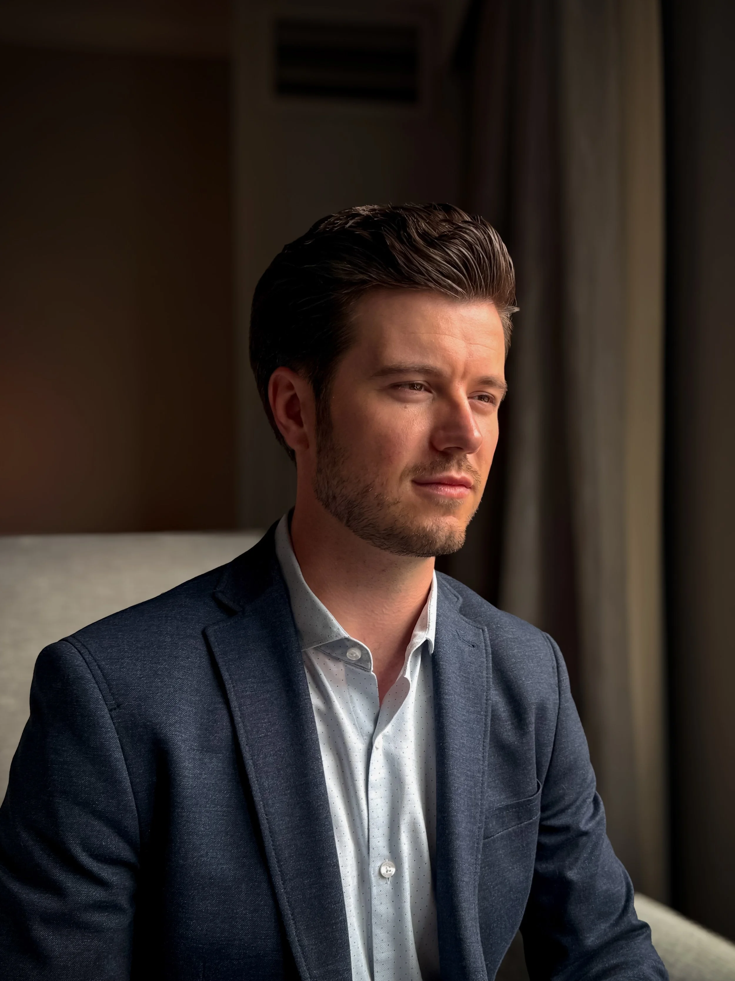 A man with dark hair and a beard wearing a dark blazer and white shirt, sitting indoors near a window, looking thoughtfully out of frame.