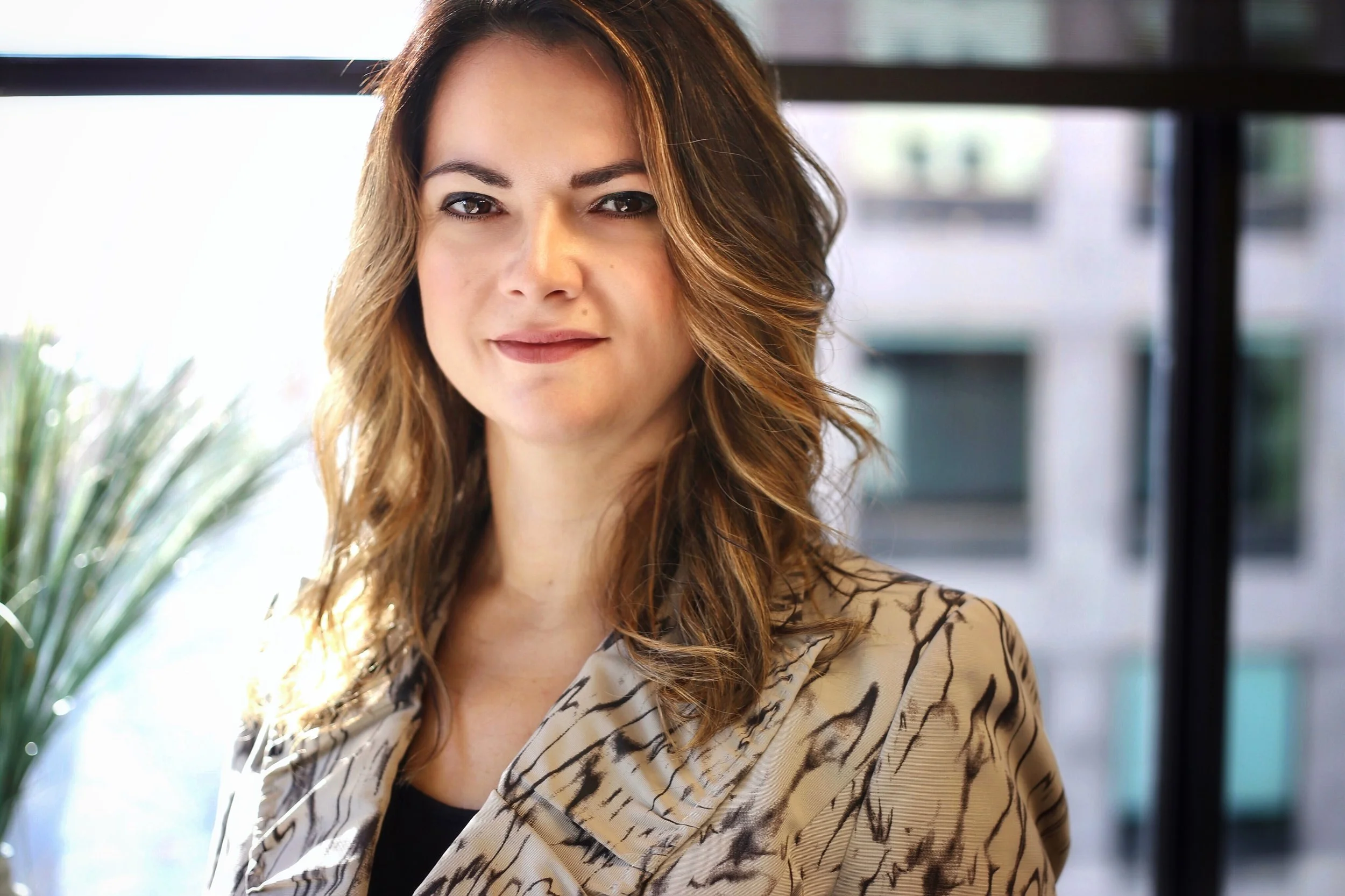 Close-up of a woman with shoulder-length brown hair and light skin, wearing a patterned beige blazer over a black top, standing indoors with a blurred cityscape background.