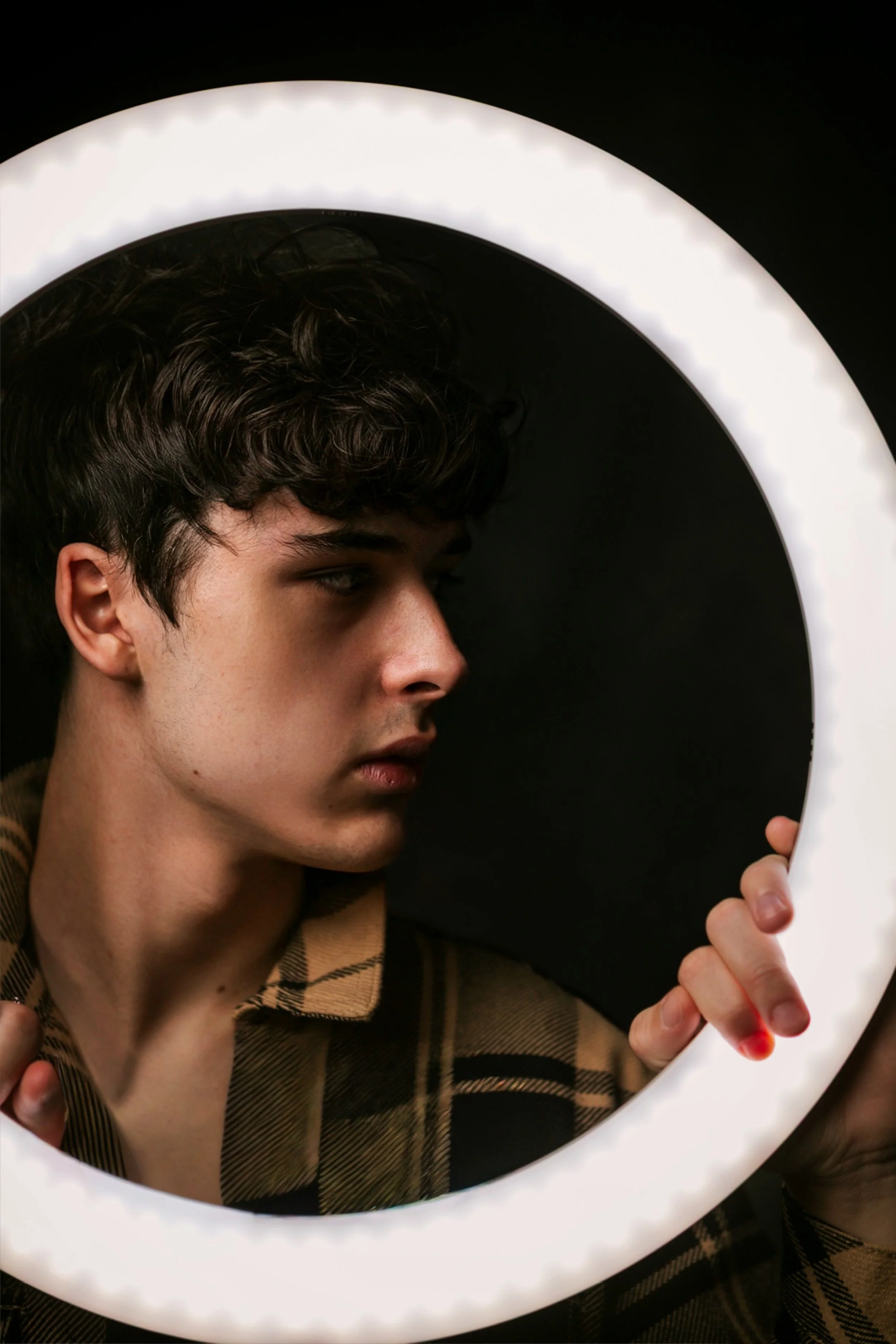 Young man with curly hair looking to the side through a circular LED mirror or light.