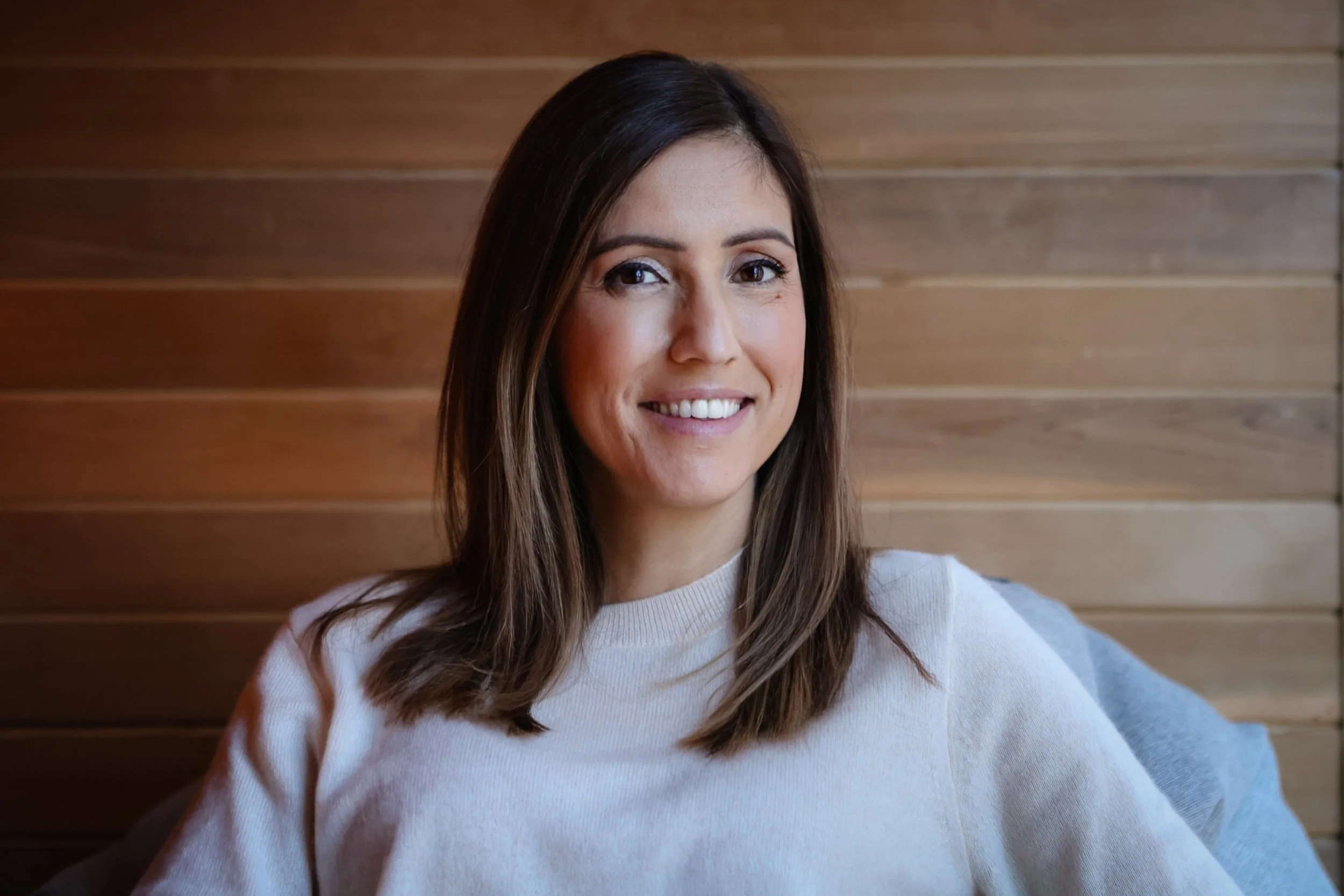 A smiling woman with shoulder-length brown hair, wearing a light-colored sweater, sitting in front of a wooden wall.