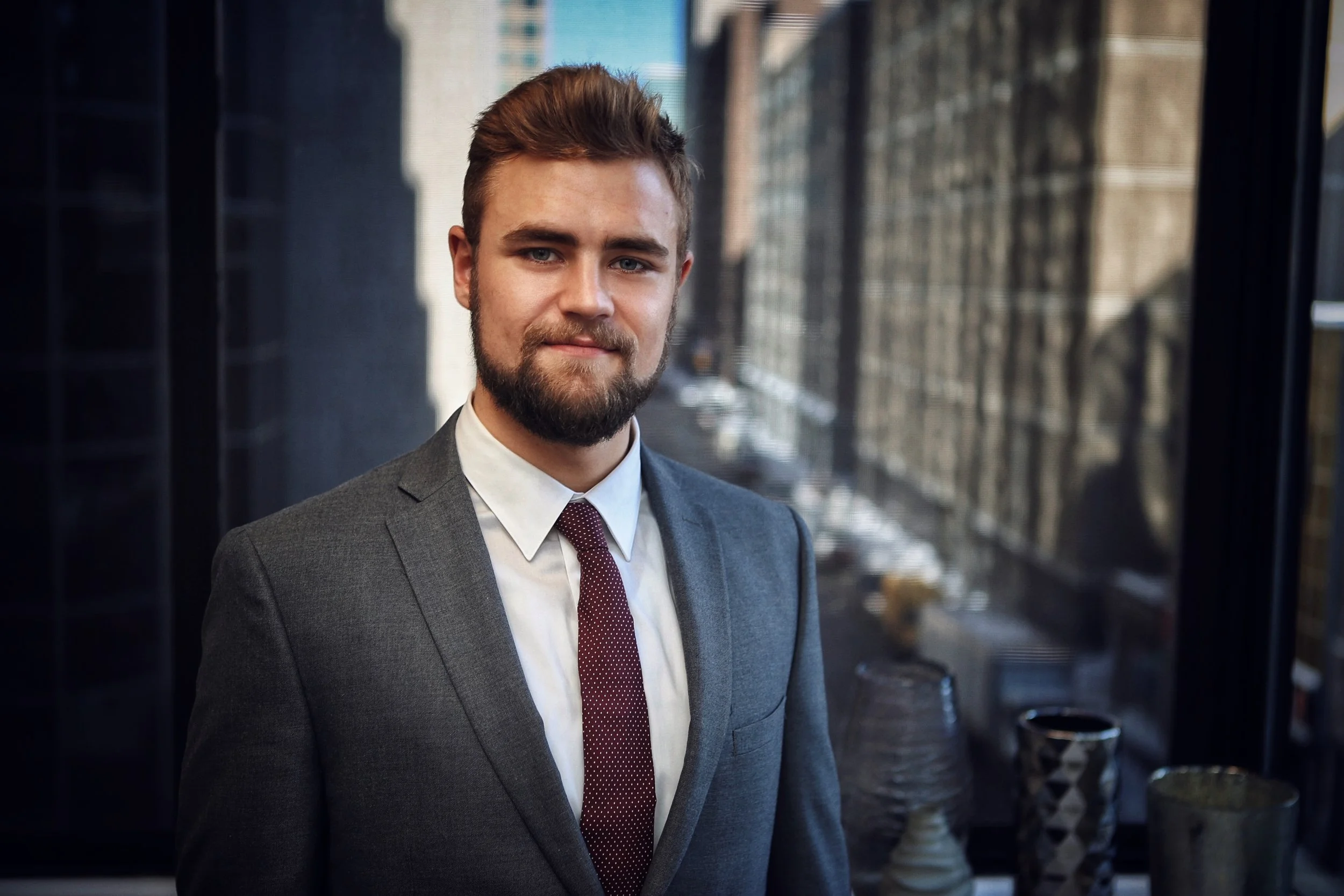 A young man with a beard wearing a gray suit, white shirt, and maroon tie, standing indoors near a large window overlooking a city street with buildings and cars.