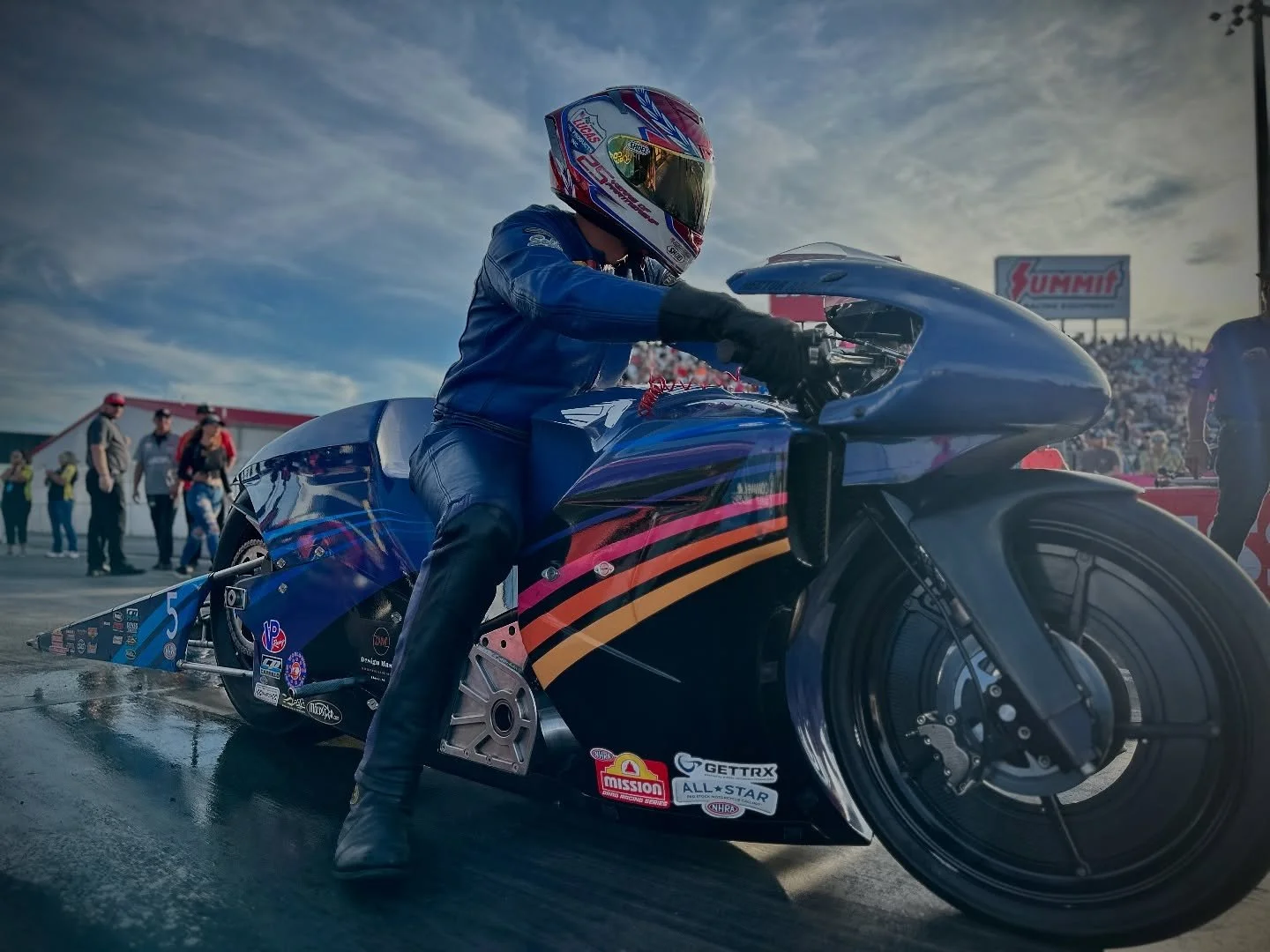 Professional motorcycle racer in blue racing suit and helmet riding a black and blue drag racing motorcycle on a race track, with a crowd in the background and a sky with clouds.