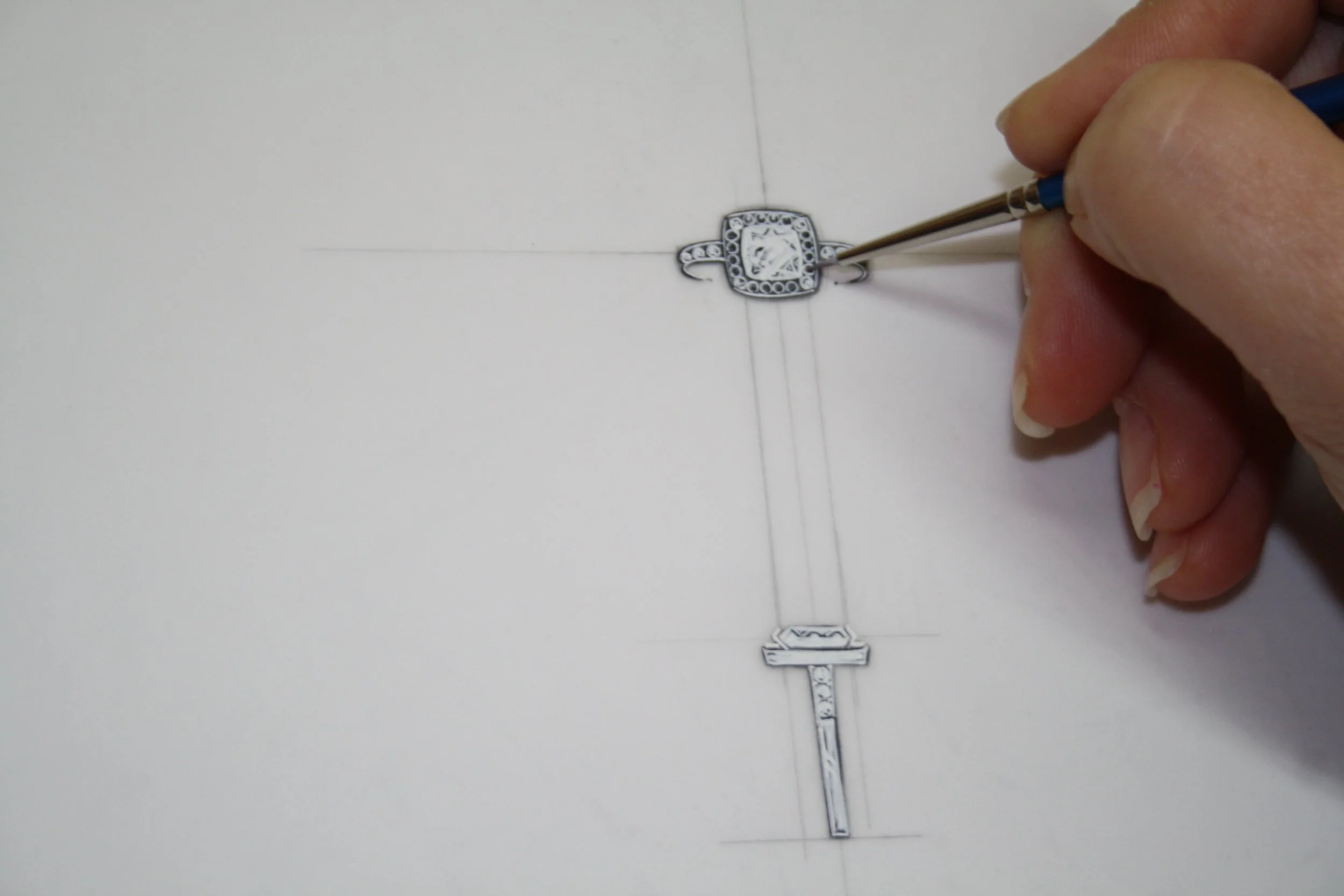 Hand holding a small precision tool working on a silver decorative piece with a square centerpiece, placed on paper with drawn lines.