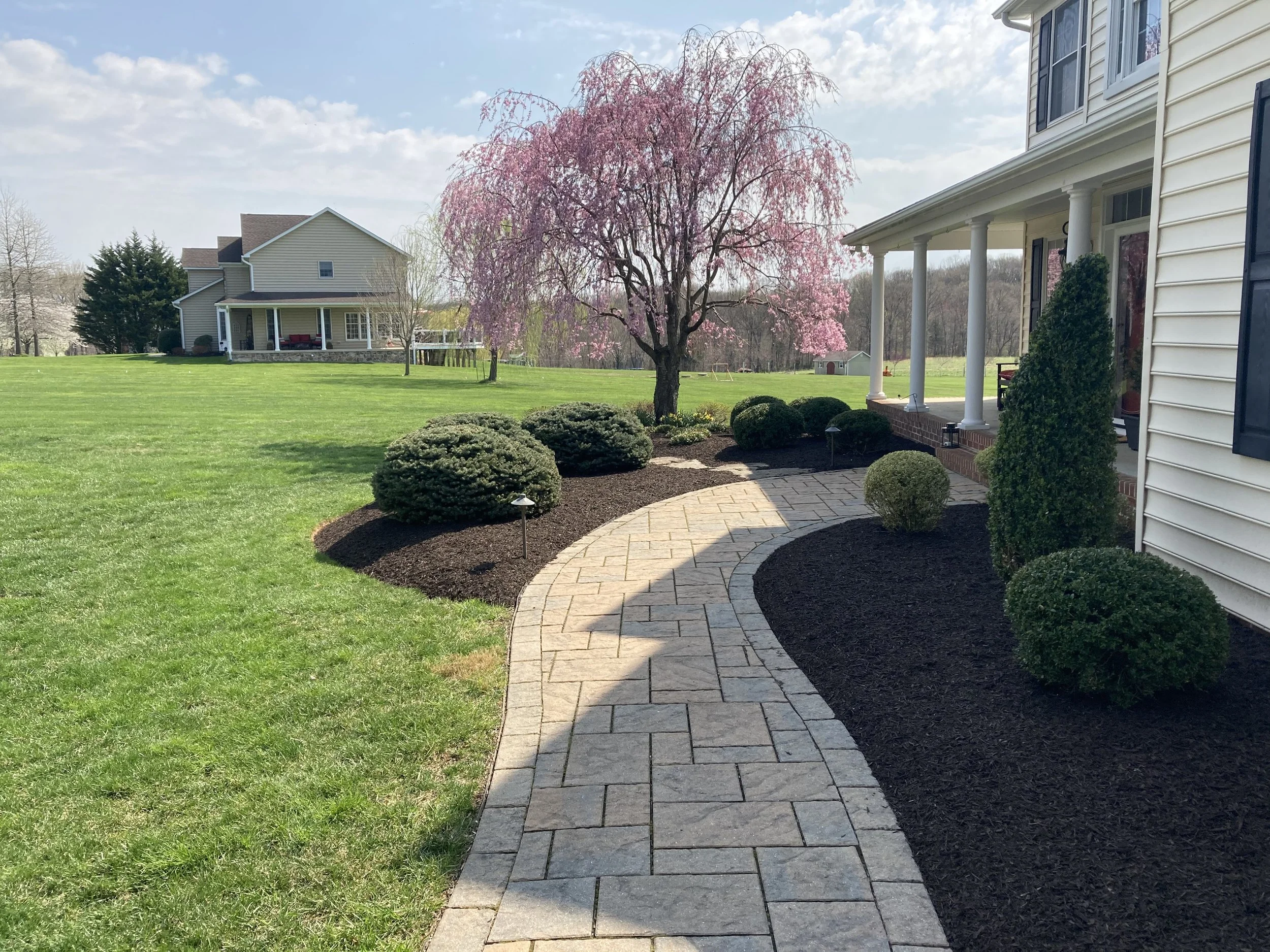 A curved stone pathway leading from a house porch to a grassy yard, with neatly trimmed bushes and a pink flowering tree in full bloom, under a partly cloudy sky.