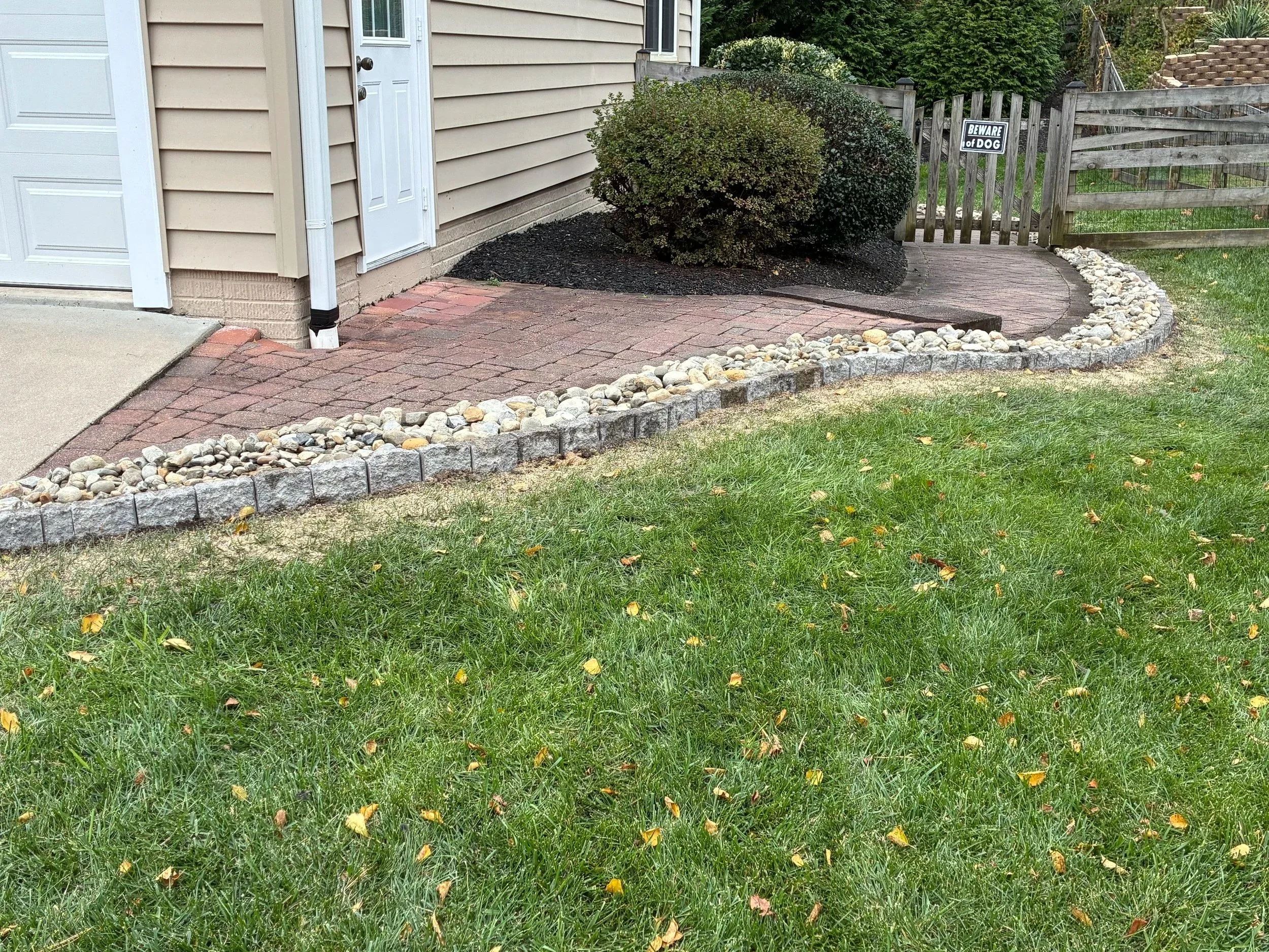 A backyard with a green grass lawn, small yellow leaves scattered on the grass, a curved stone and brick pathway leading to a wooden gate, and a bush near the house with a sign that says 'Beware of Dog'.