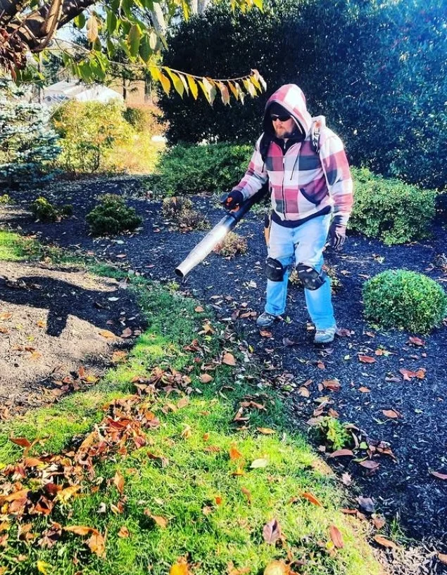 Person using leaf blower in a garden with green grass, bushes, and trees, wearing a checkered hoodie, white pants, and safety gear.