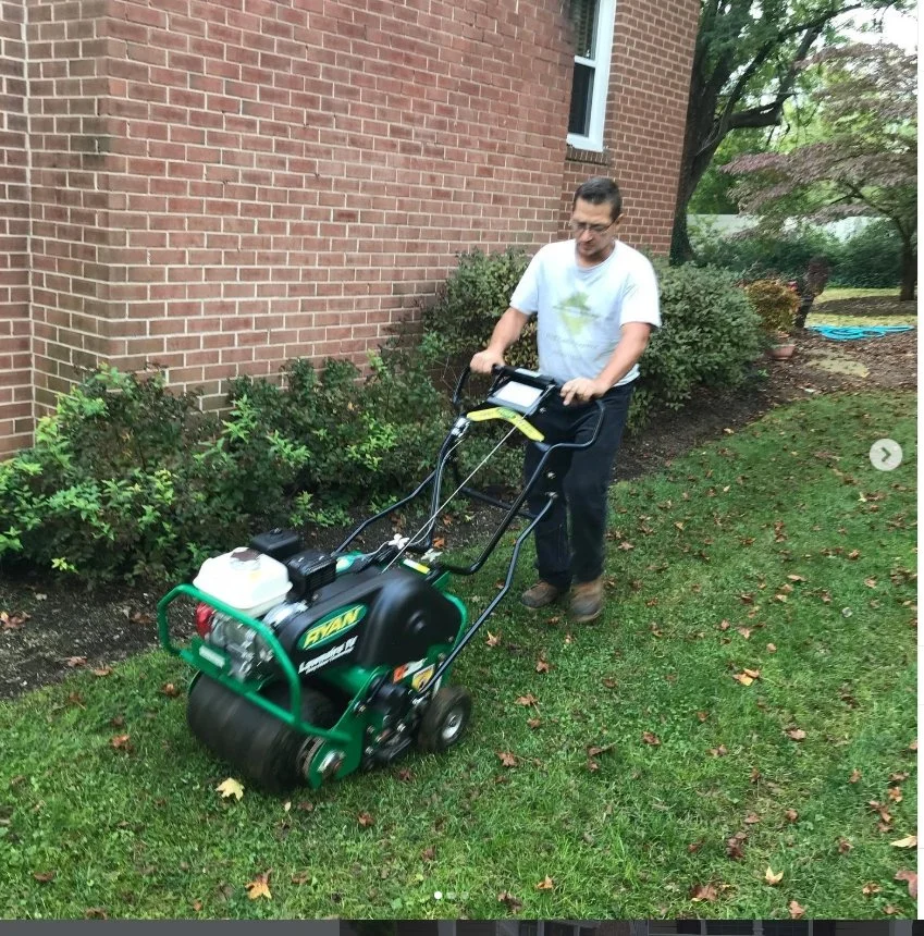 A man operating a small green and black lawn roller on a grassy area next to a brick house, with bushes and trees in the background.