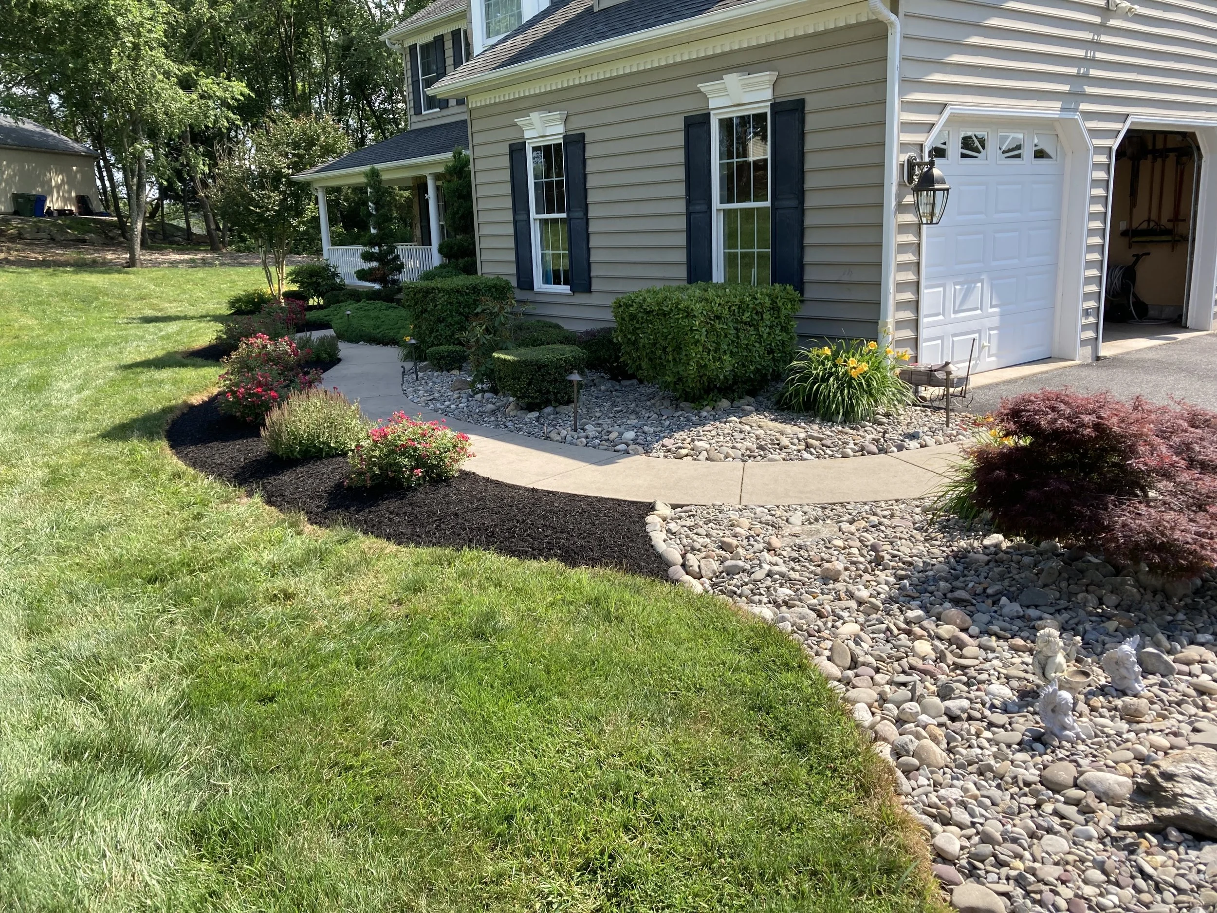Photo of a well-maintained front yard with a curved walkway, landscaped garden beds with colorful flowers, bushes, and decorative stones, adjacent to a beige house with black shutters and a garage door.