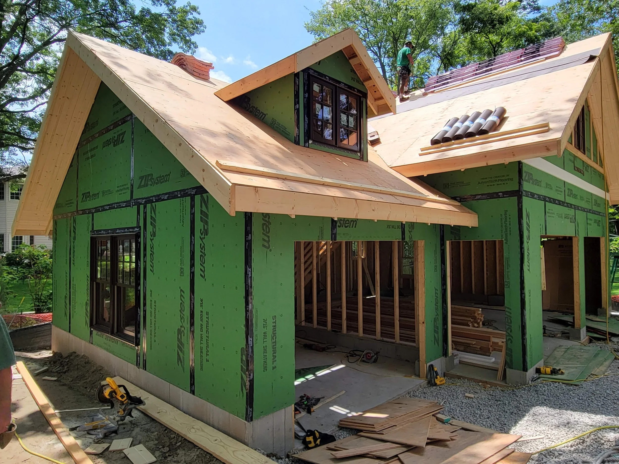 A house under construction with green sheathing and a wooden roof. The roof has multiple sections and a small window. A construction worker is working on the roof, and various tools and building materials are scattered around.