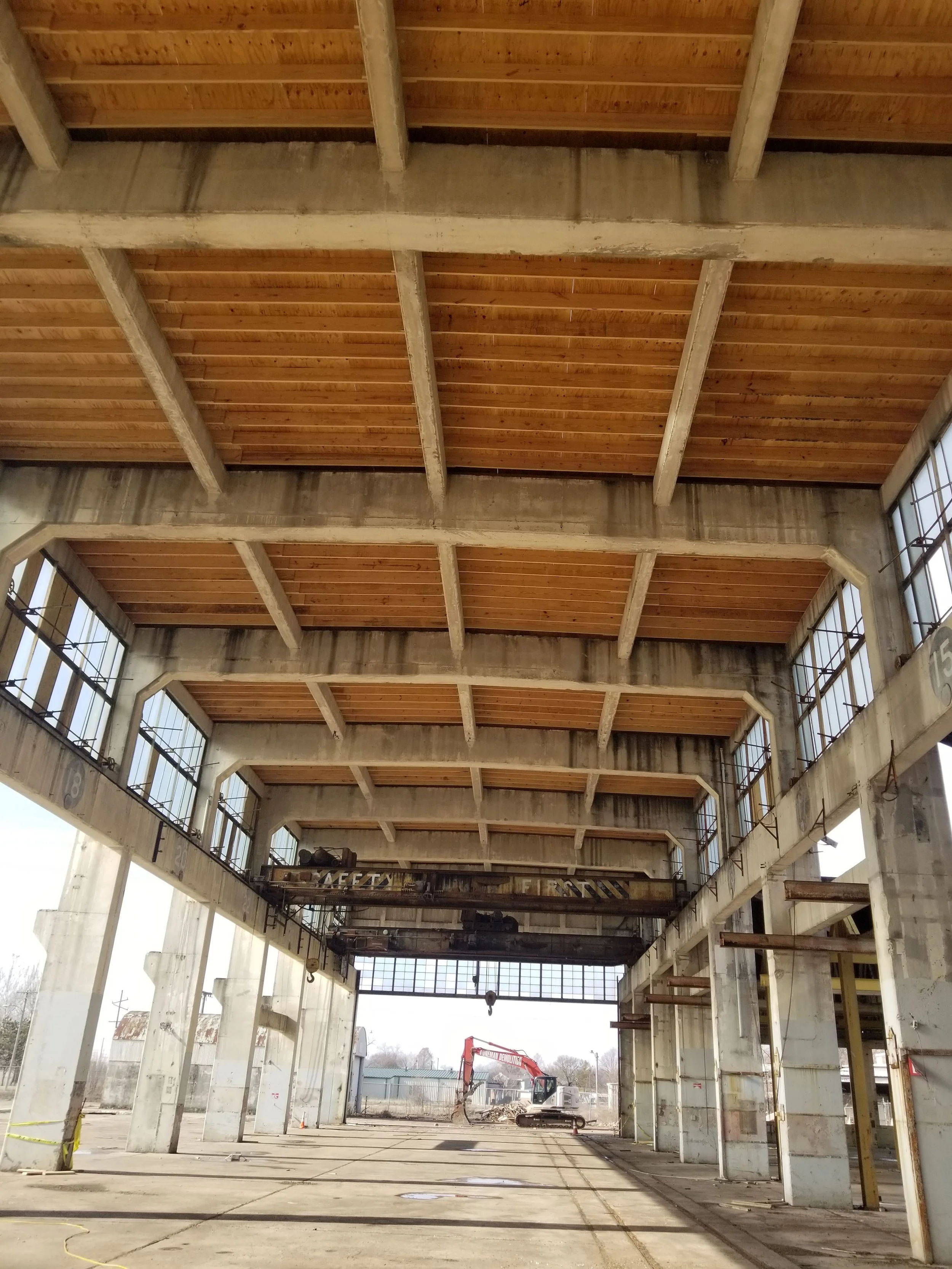 Interior of an industrial building under construction with a high wooden ceiling, concrete beams, large windows, and construction equipment inside.