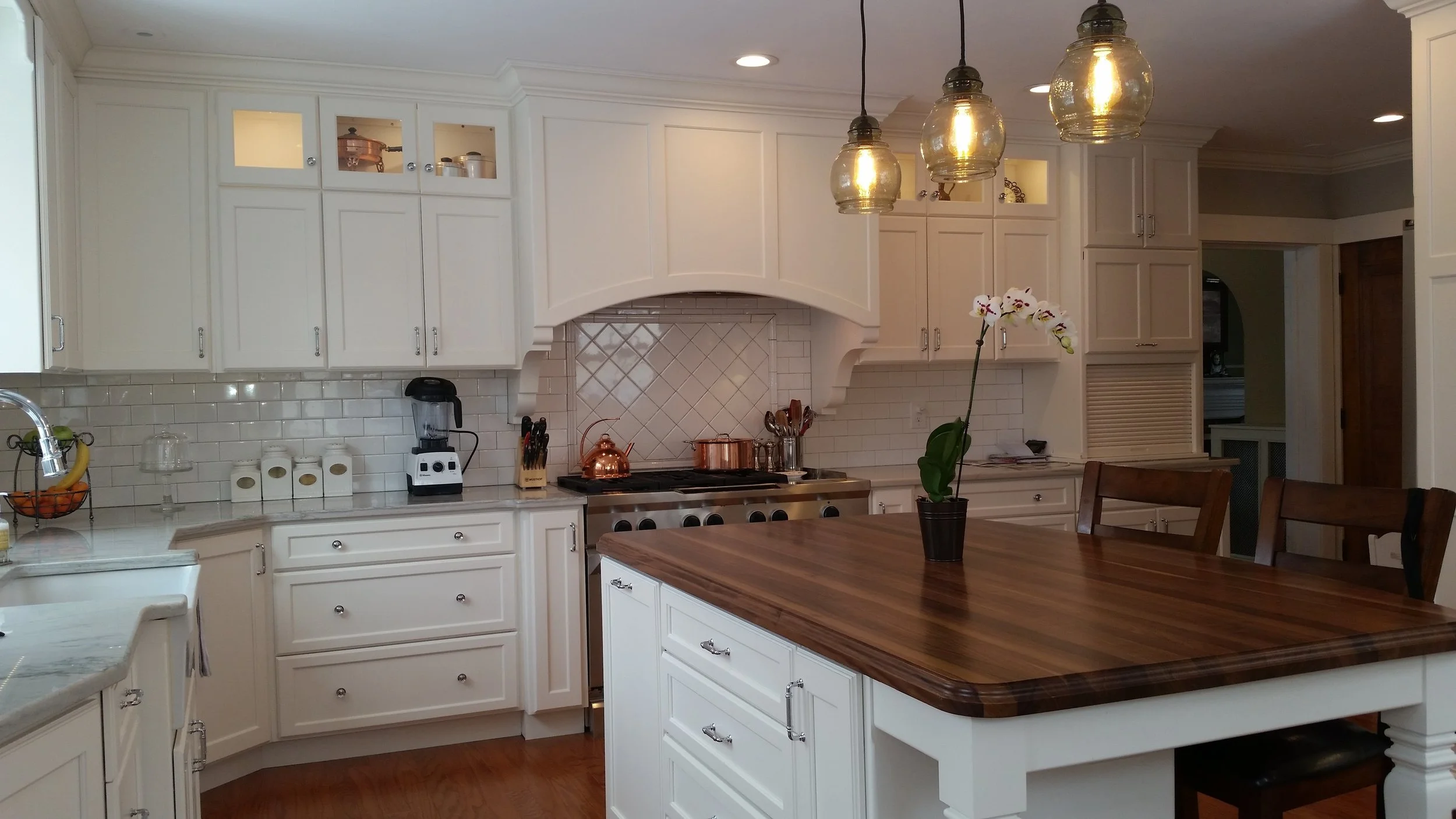 White kitchen with wooden island, orchid on table, hanging pendant lights, and various appliances and utensils on countertops.
