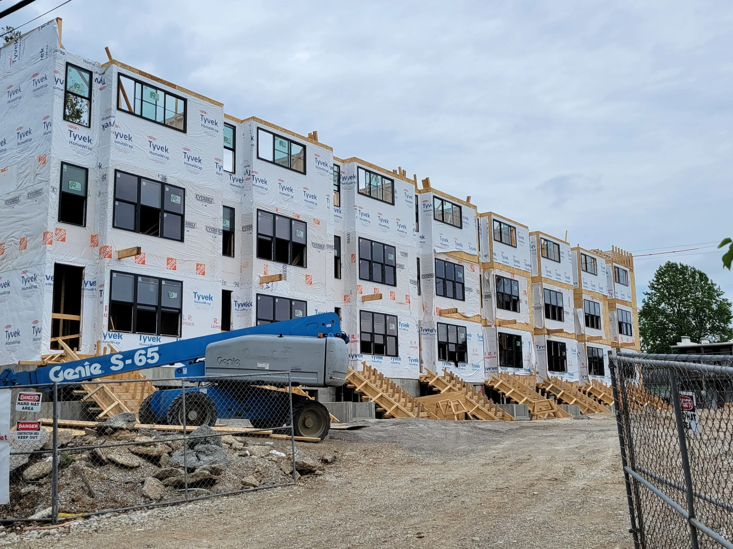 Construction site of a multi-story building with exterior walls covered in Tyvek HomeWrap, large black-framed windows, and wooden stairs leading to entrances, with a Genie S-65 lift and a chain-link fence in the foreground.