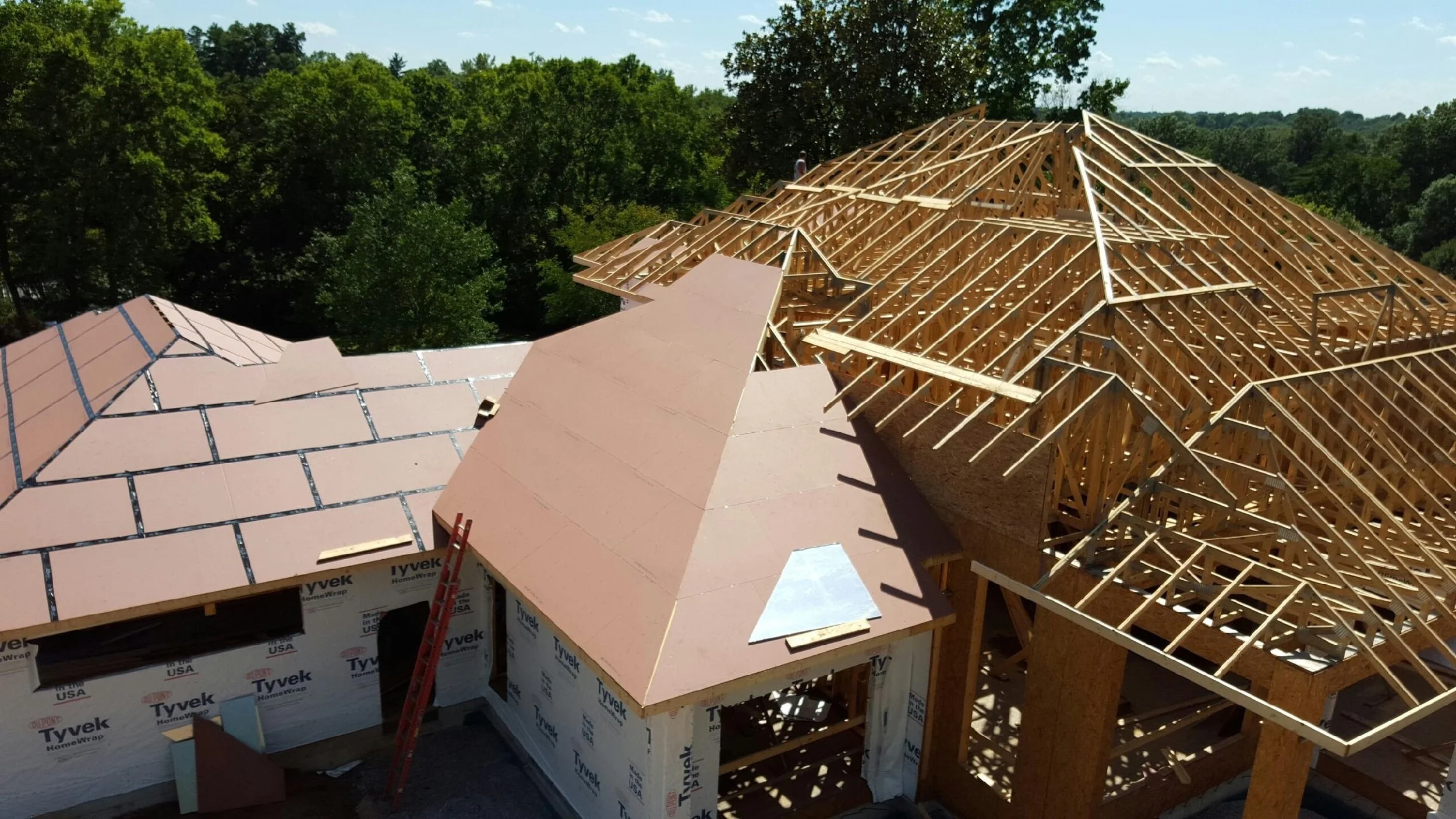 A house under construction with a partially completed roof. The framework is visible with wooden beams and trusses, while some sections have roofing material installed.