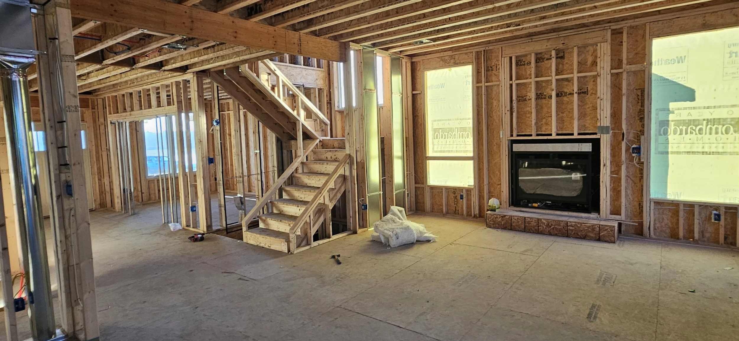 Interior of a house under construction with exposed wood framing, staircase, and a fireplace opening. Windows are covered with yellow building wrap, and construction materials and tools are scattered on the floor.