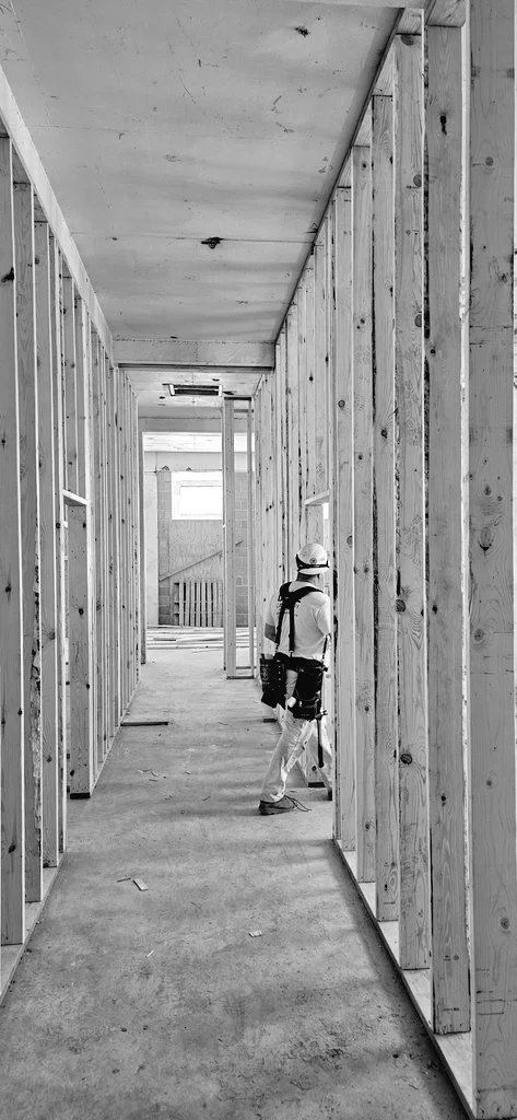 A construction worker standing in a partially built indoor corridor with wooden studs and an unfinished ceiling.