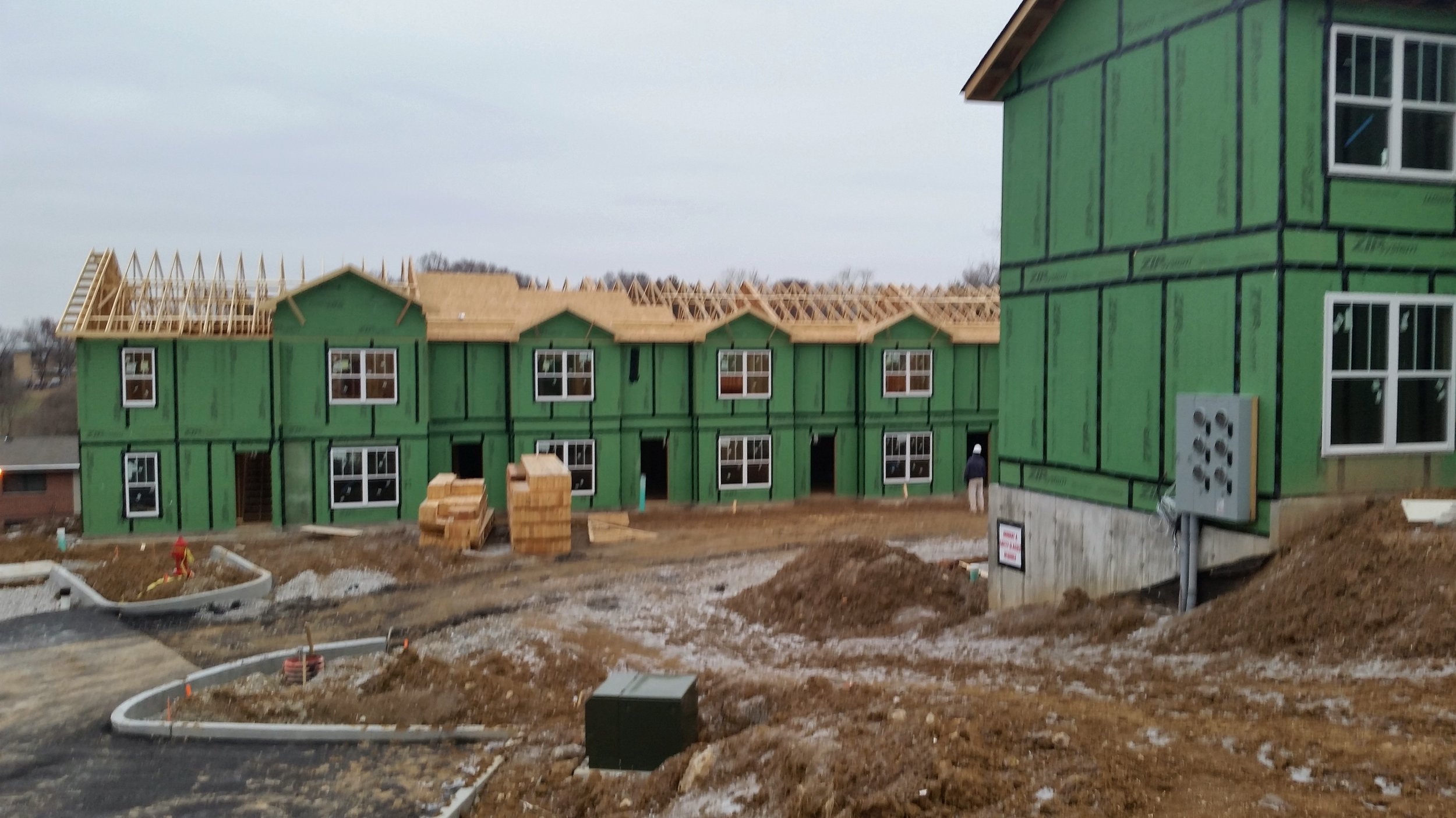 Multiple townhouses under construction with green sheathing and some roofs partially built, construction materials scattered on the ground, and a worker visible in the background.