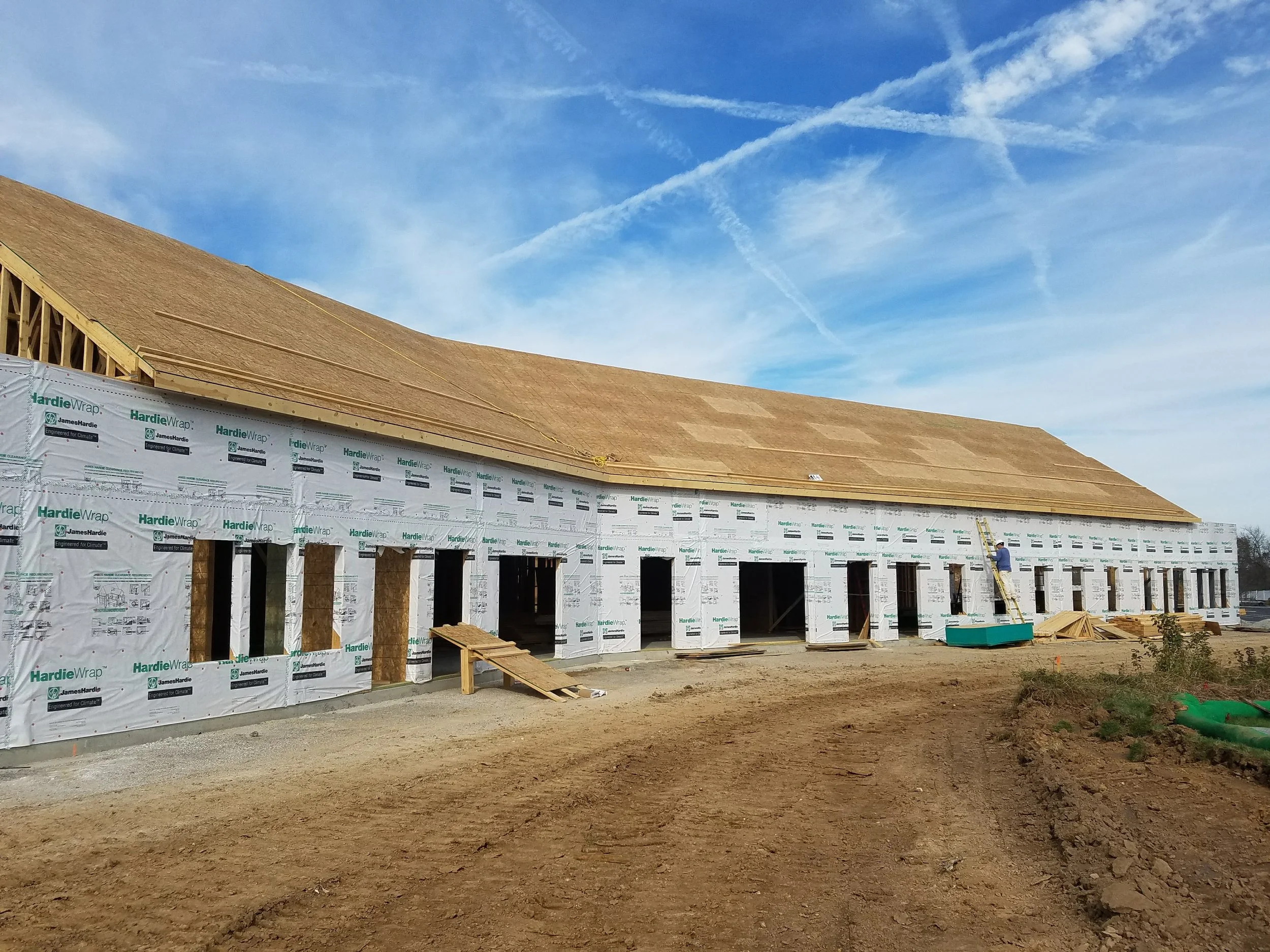 Construction site of a large building with a wooden roof and wrapped exterior walls, dirt ground in the foreground, a ladder leaning against the building, and a worker standing on the ladder, with a blue sky and contrails in the background.