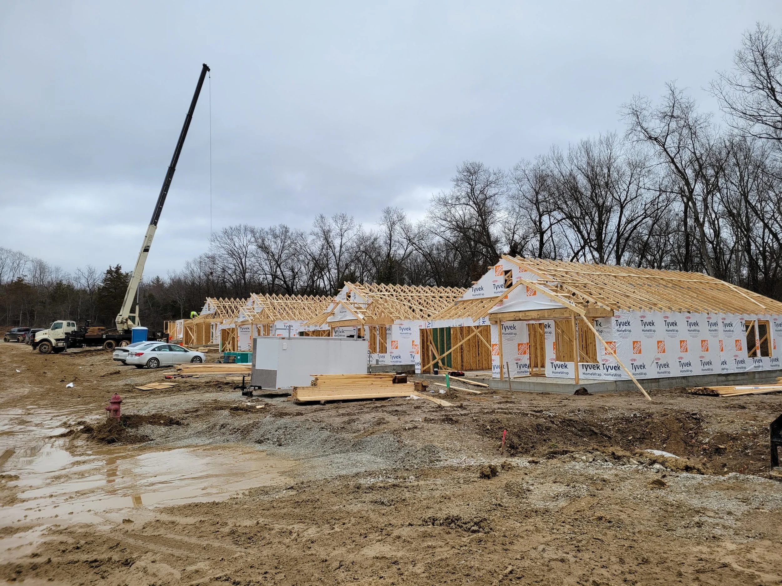 Construction site with partially built houses, a crane lifting materials, trucks, and parked cars on muddy ground, with leafless trees in the background under a cloudy sky.