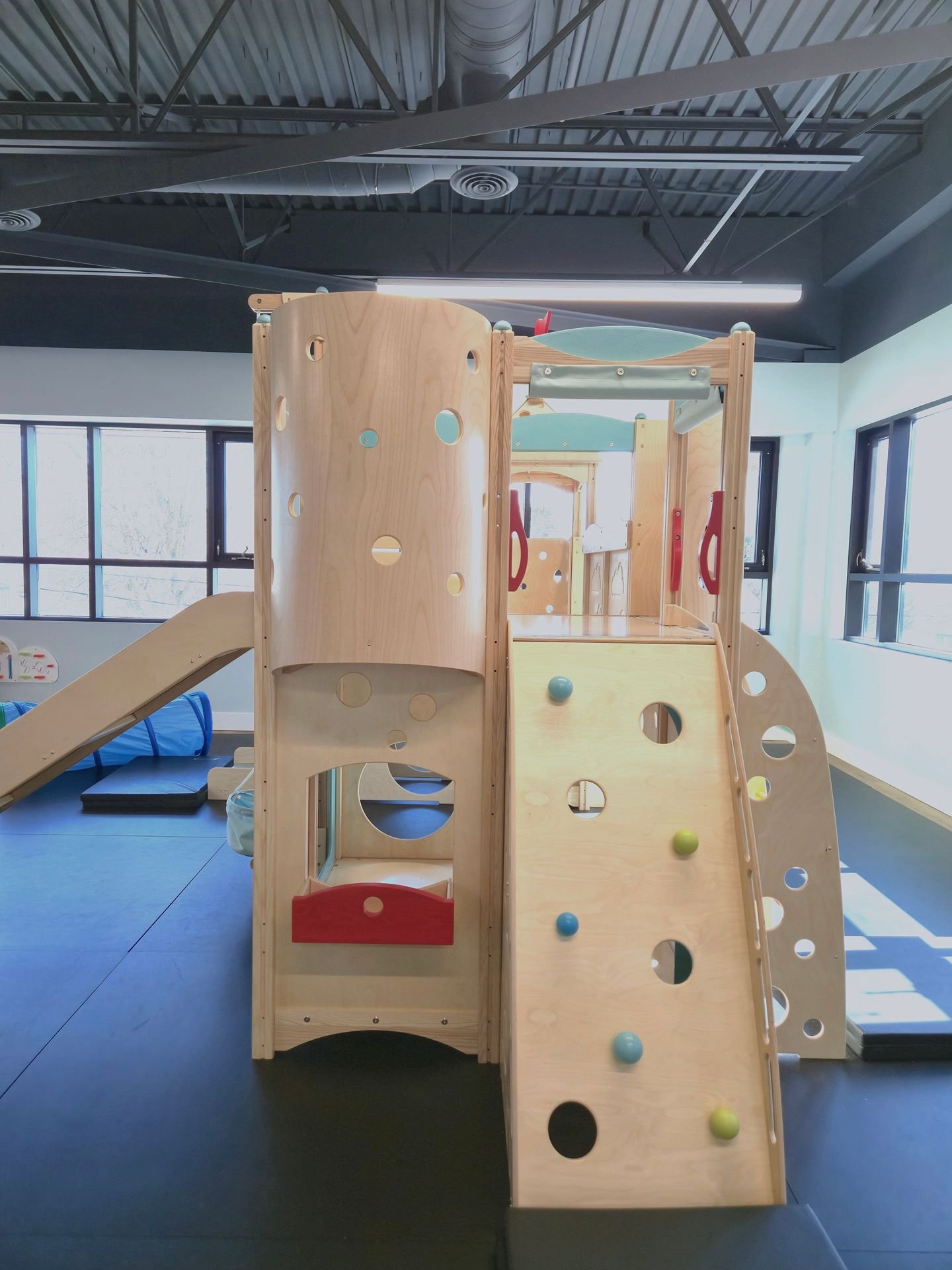 Indoor wooden playground structure with a slide, climbing wall with colored balls, and various openings, set in a room with large windows and a black ceiling.