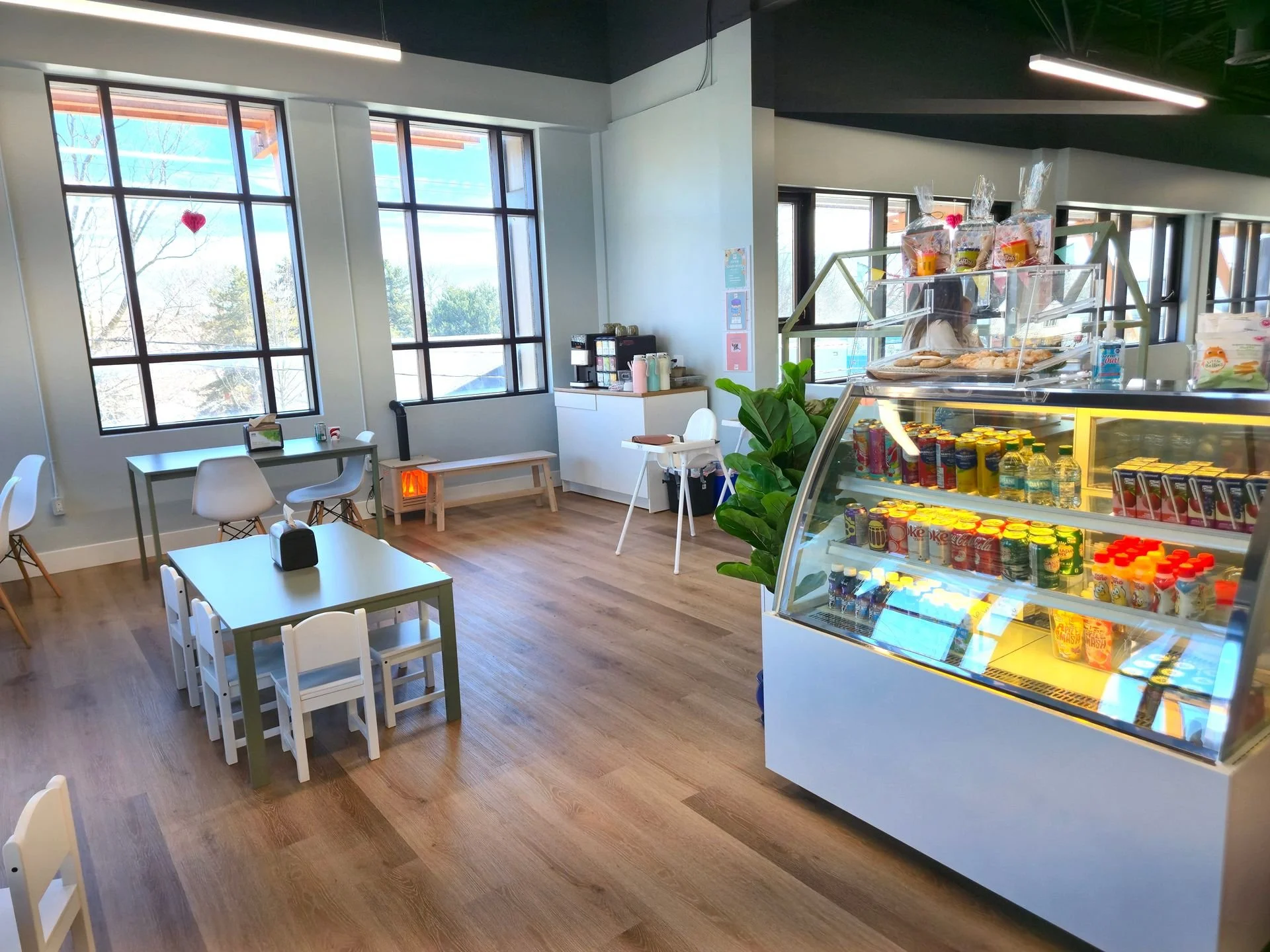 Interior of a cozy cafe with large windows, several small tables with chairs, a refrigerated display case with assorted bottled drinks, a snack shelf on top, and a coffee station in the corner.