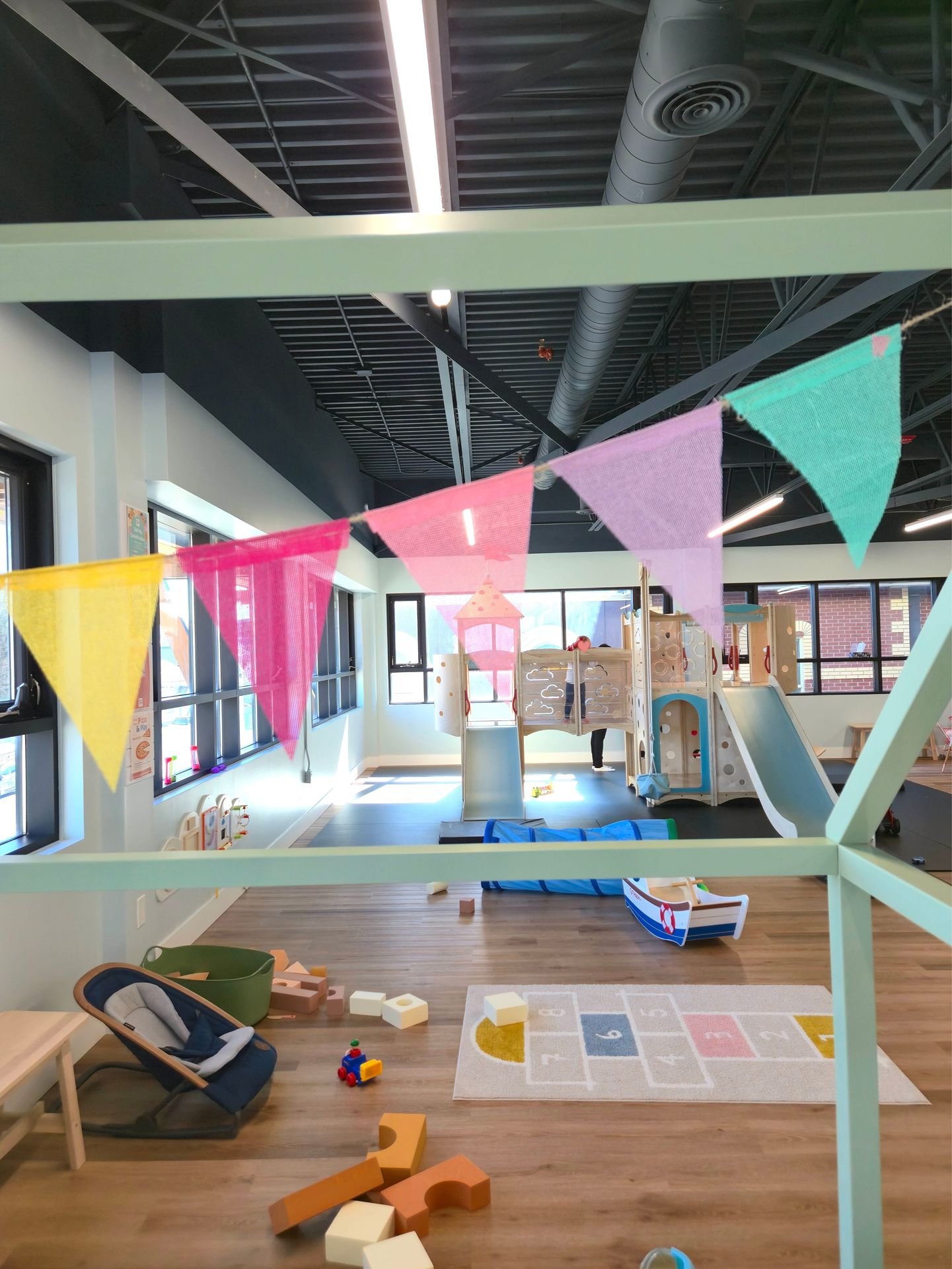 Indoor children's play area with a foam climbing structure with slide, toy boat, hopscotch rug, scattered toys, and colorful pennant banners hanging overhead.