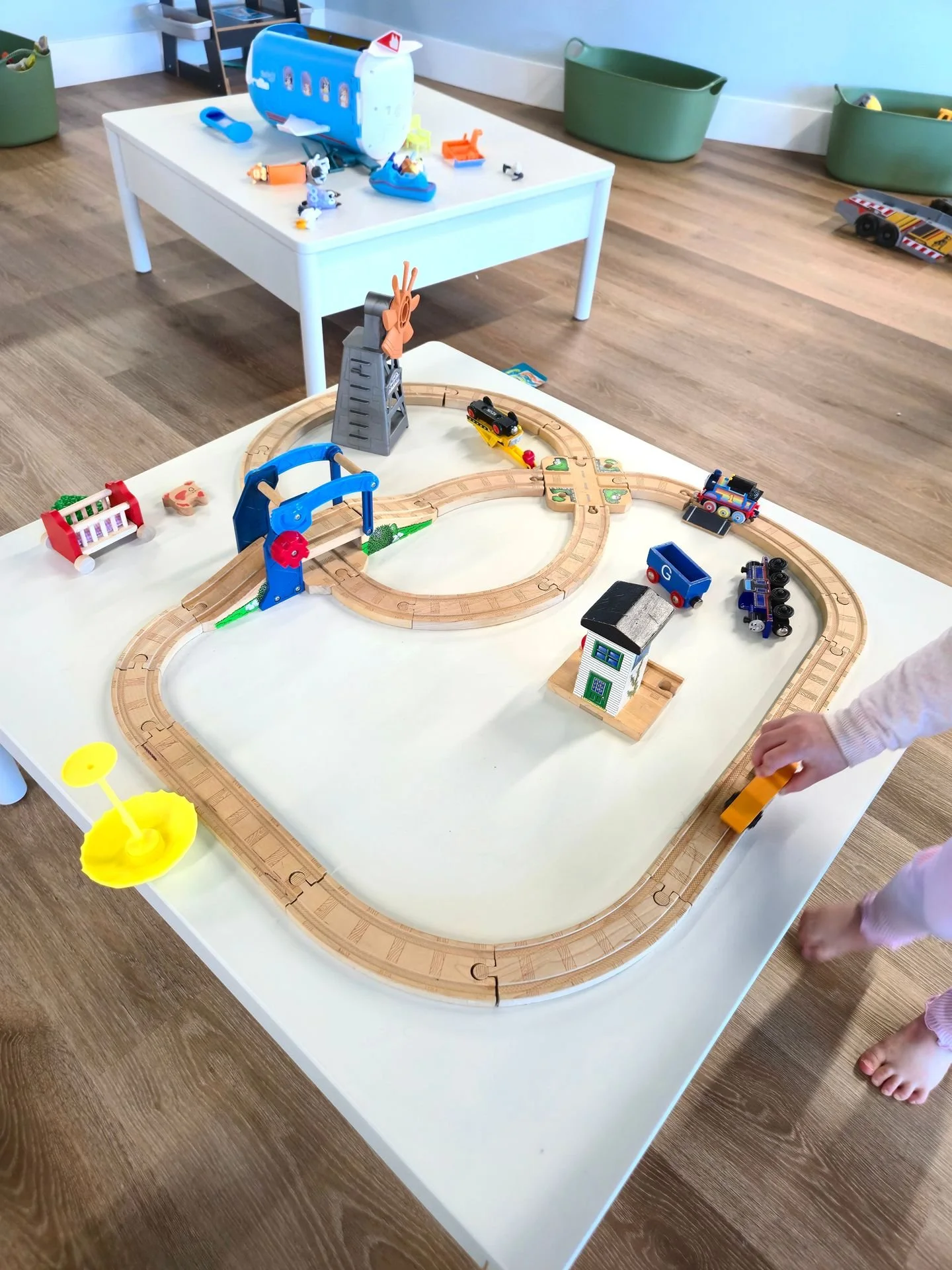 Children playing with a wooden train set on a white table, featuring tracks, toy trains, a small house, and accessories in a playroom.