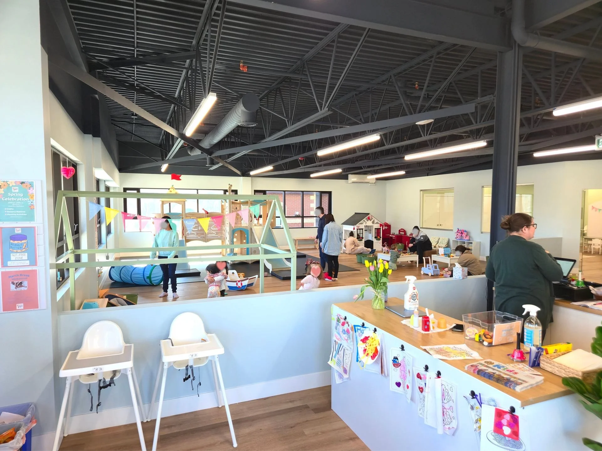 An indoor playroom with children playing and adults supervising. There are toys, a playhouse, a tunnel, and a slide. Front area has a counter with a vase of flowers, sanitizer, and children's drawings displayed. The ceiling has exposed dark industria