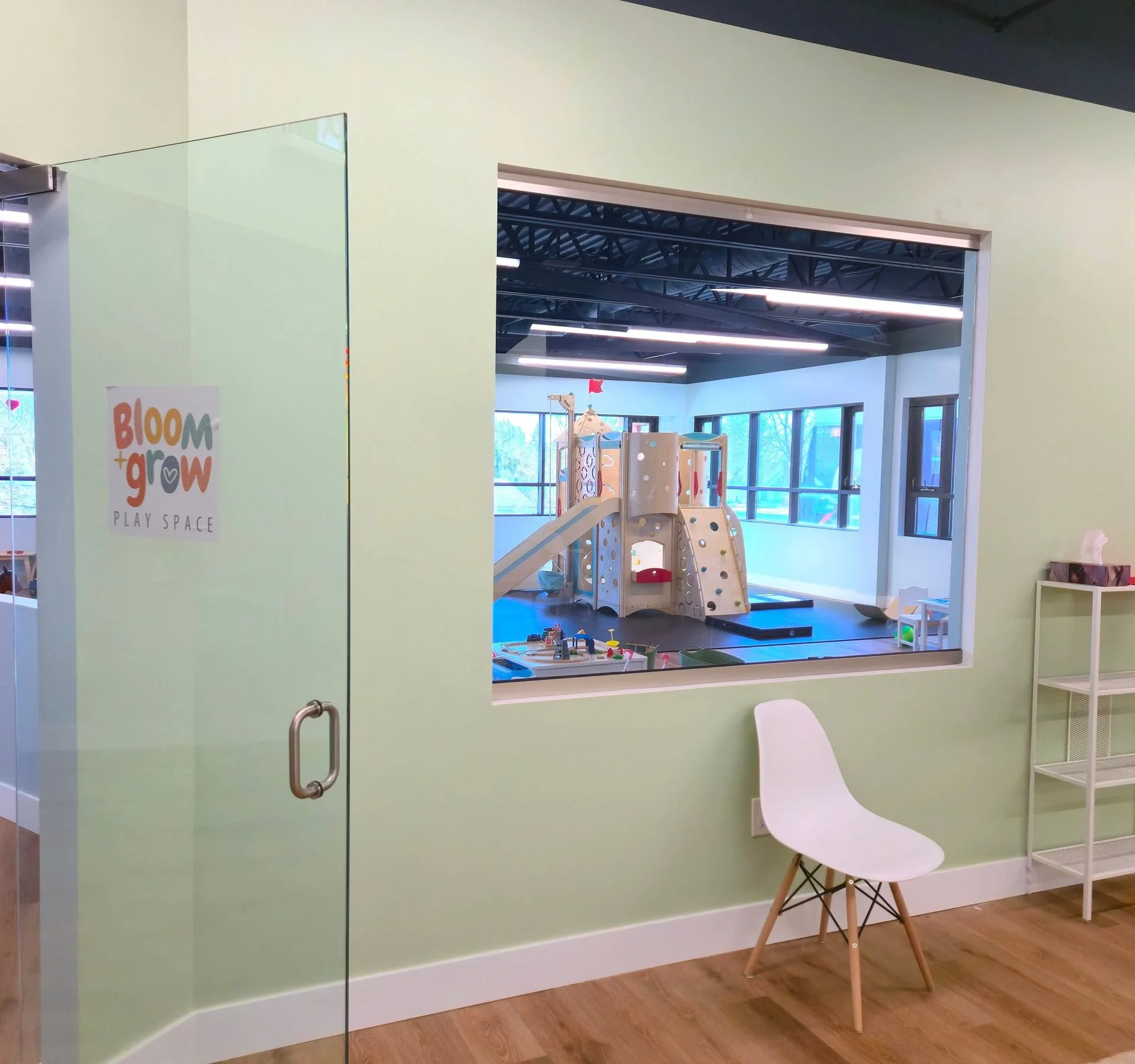 View of an indoor children's play area seen through a large window in a green-walled room. The play area includes a small slide and climbing structure, with bright natural light from windows and a black ceiling.