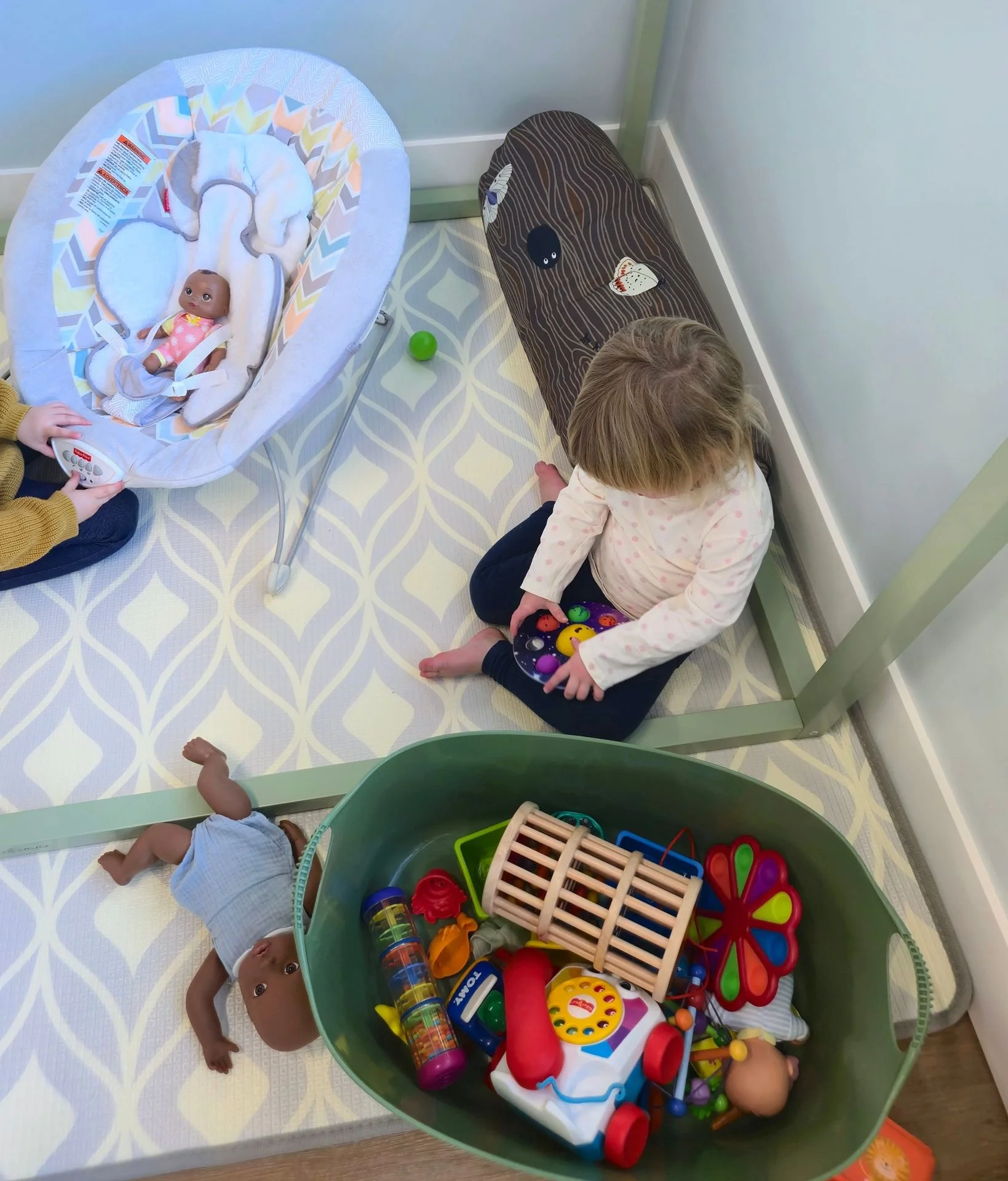 Child sitting on the floor near a green toy storage bin filled with various toys, including a baby doll, plastic animals, and a toy stroller, with a doll in a bassinet nearby.