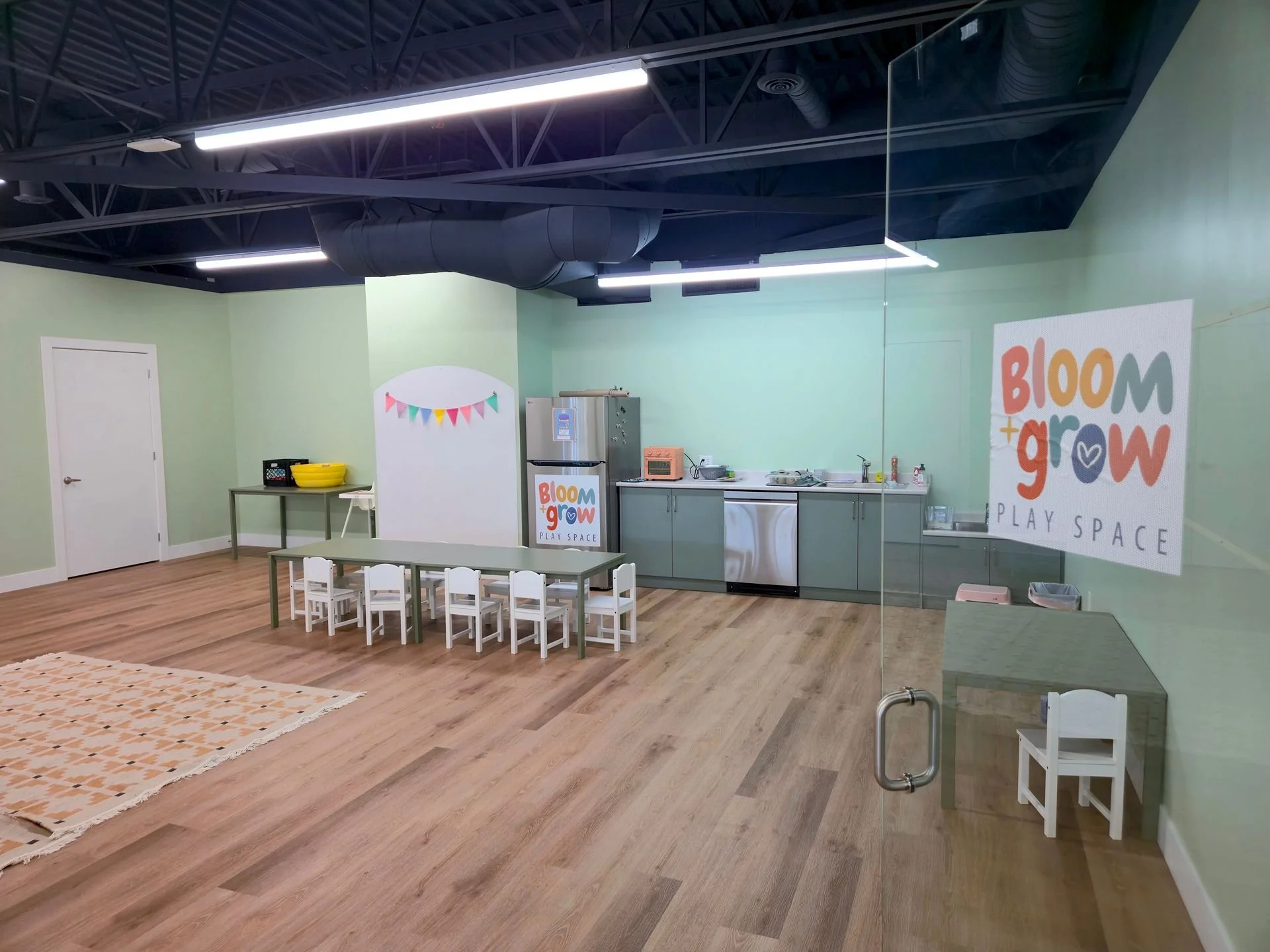 Children's play area with a small kitchen setup, table and chairs, and a decorative bunting banner, inside a room with light green walls and wood flooring, sign reading 'Bloom + Grow Play Space' on the wall.