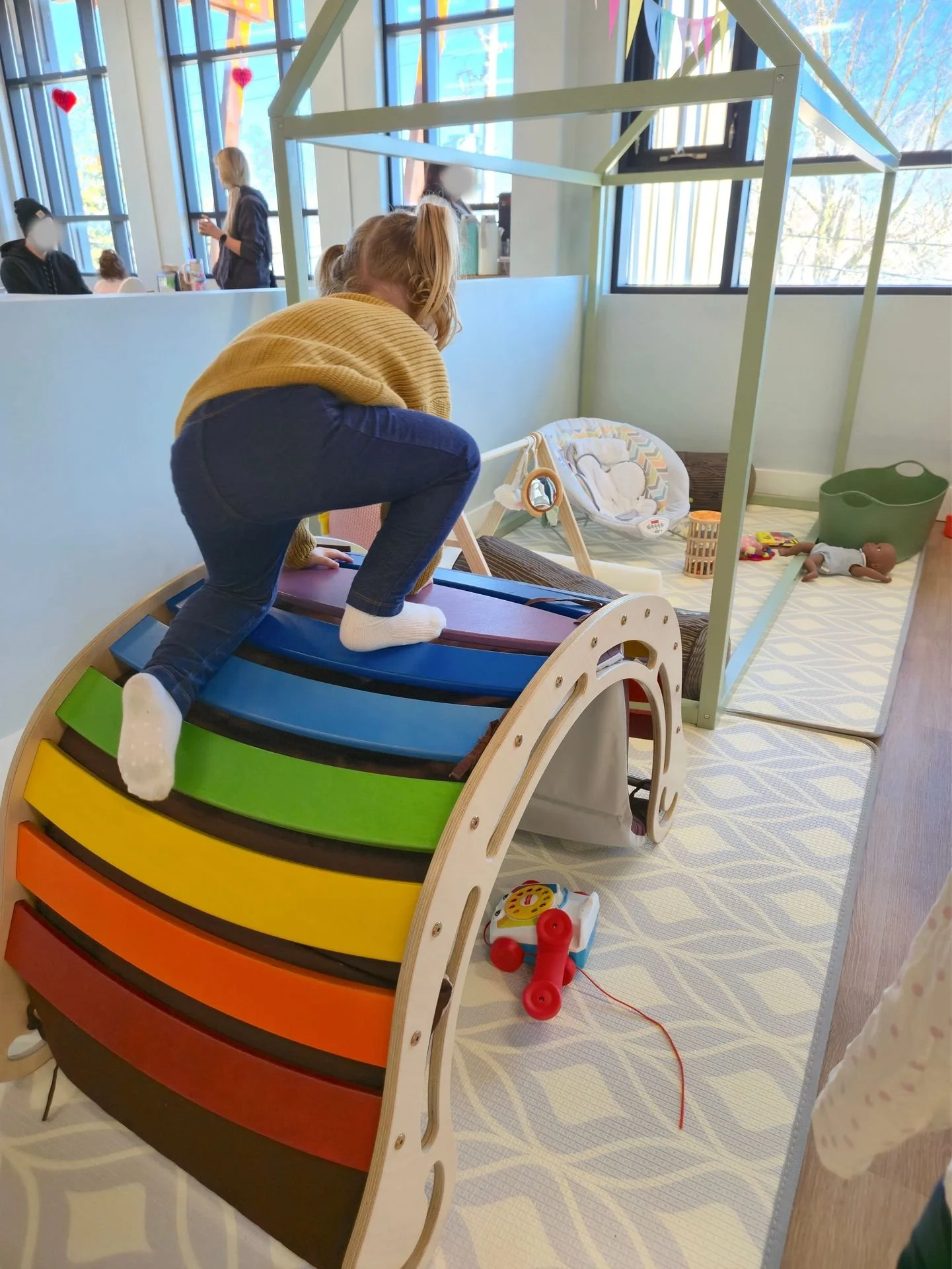 A child climbing a rainbow-colored wooden ramp in a play area at a daycare or play center, with toys and a baby doll nearby.