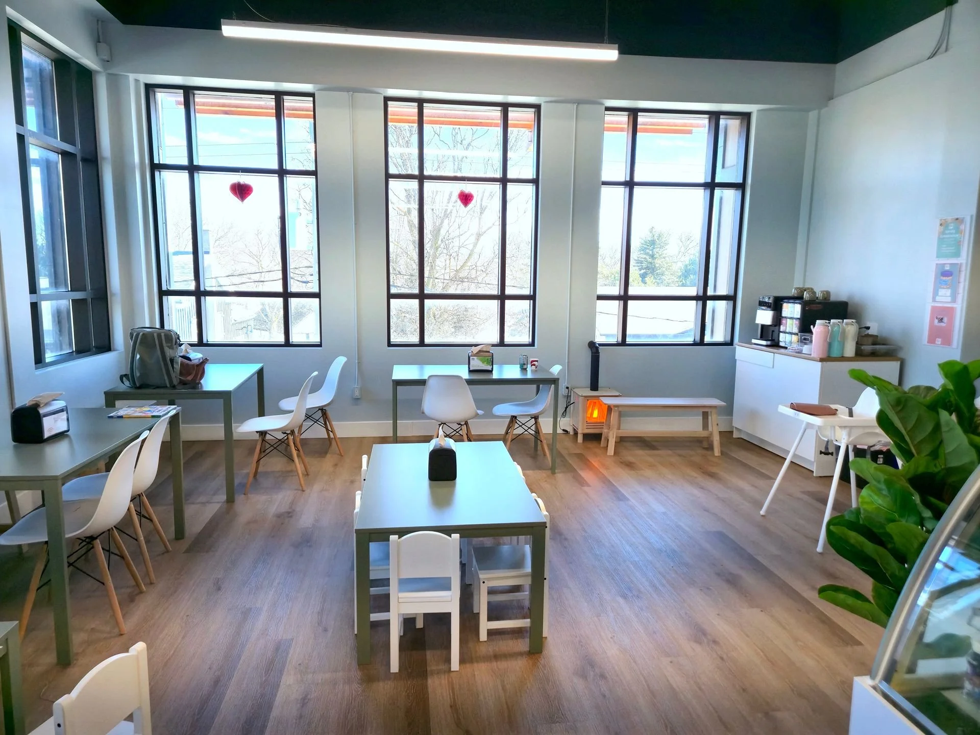 Bright cafe or lounge with large windows, white tables, and white chairs. Heart decorations hang in the windows, and there is a plant in the foreground. A heater or stove is visible near the back.