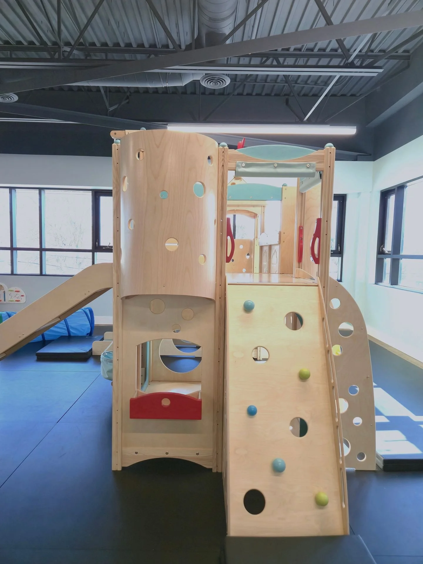 Indoor wooden play structure with slide, climbing wall, and various holes for children to play.