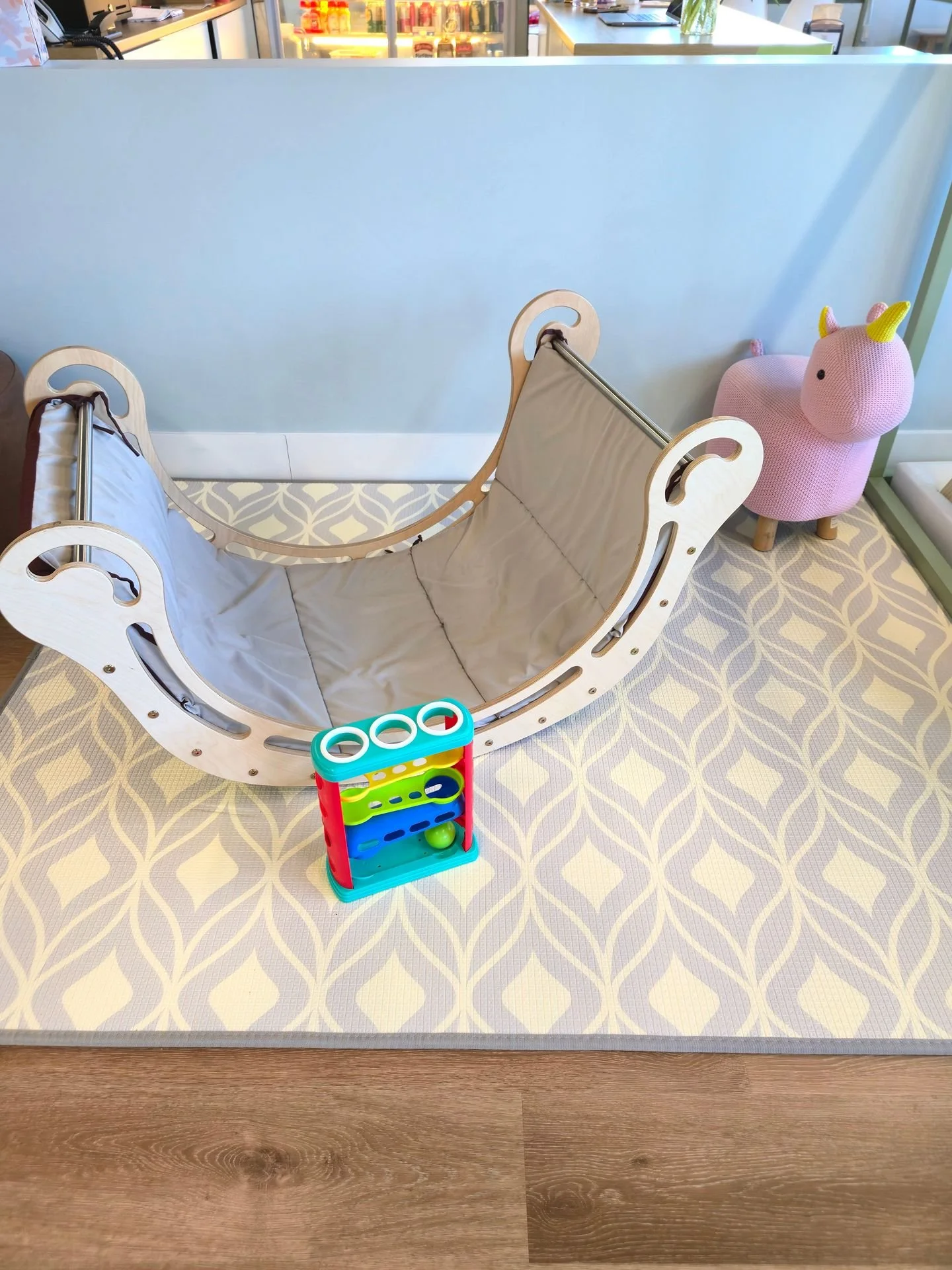 A children's play area with a light-colored wooden hammock-style bed, a pink knitted unicorn stuffed animal, and a colorful ball and maze toy on a patterned mat. The background features a white partition and a display with bottles and containers.