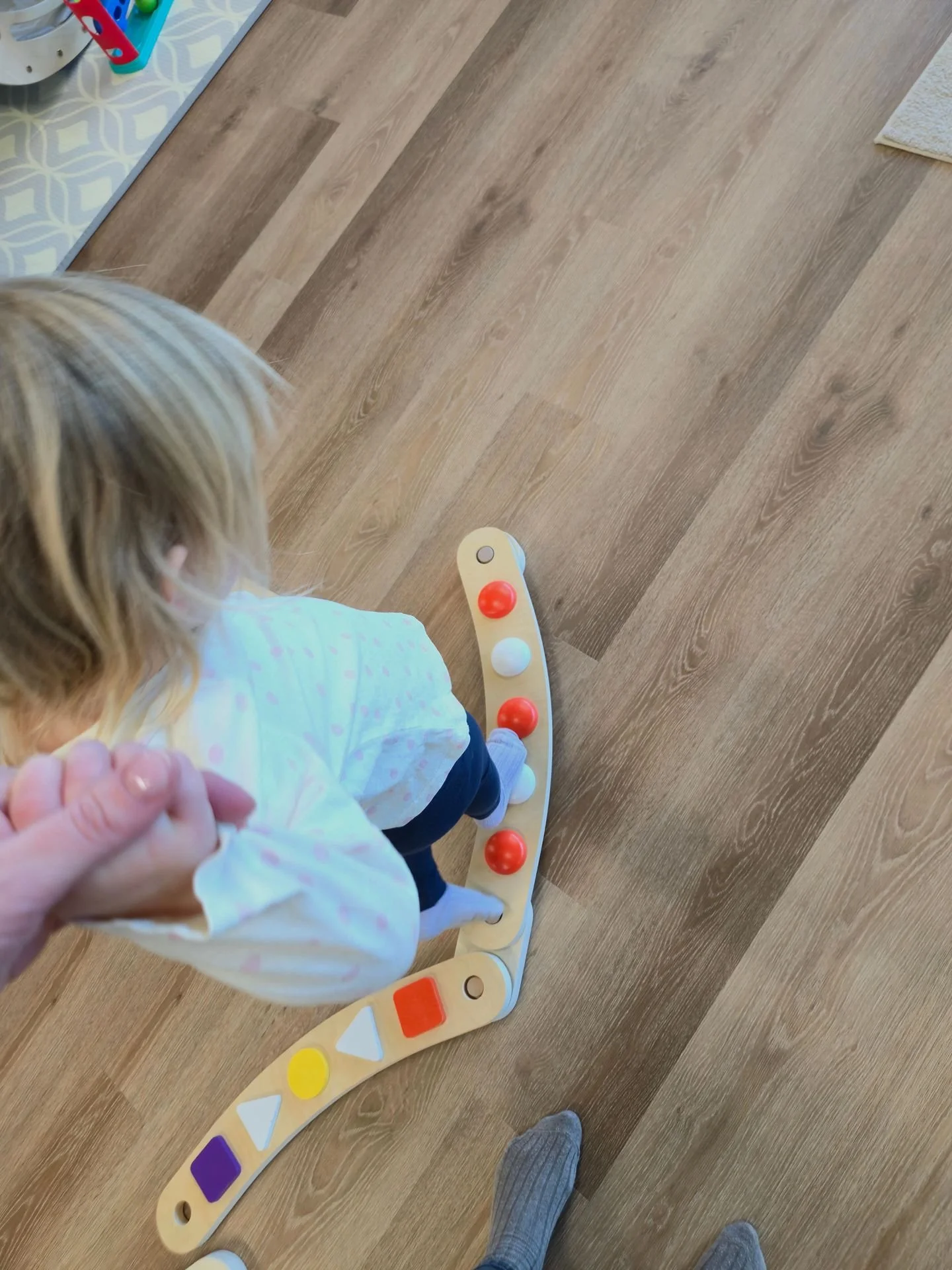 Child riding a wooden balance board with colorful geometric shapes on a hardwood floor, holding hands with an adult.