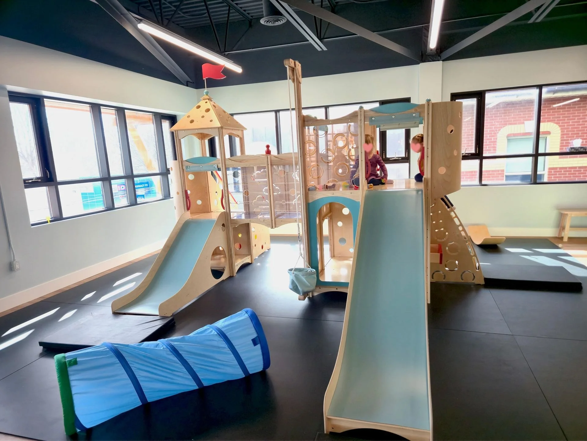 Indoor play area with a wooden playground structure featuring slides, climbing elements, a swing, and a small tower with a red flag. There are children playing, with large windows and a black ceiling with visible beams, and black flooring.