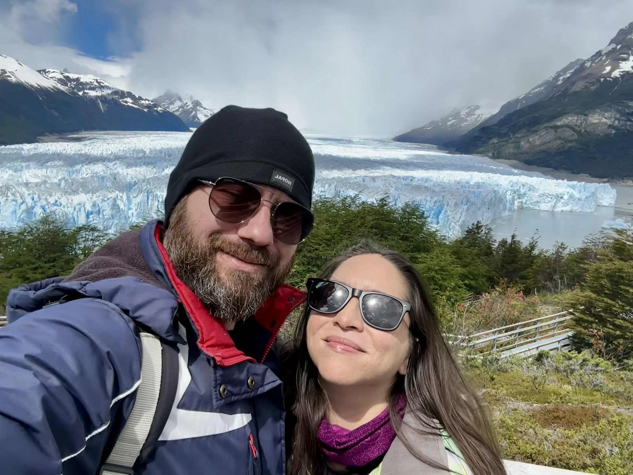 A man and woman taking a selfie outdoors with a glacier, mountains, and cloudy sky in the background.