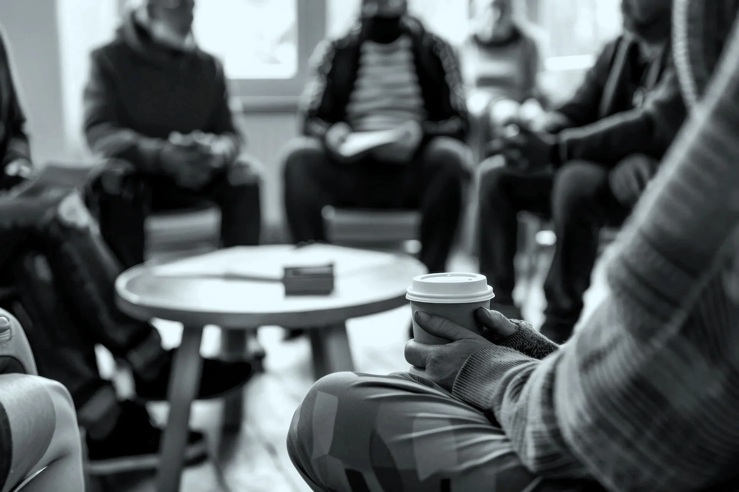 People sitting in a circle in a room, some holding papers or phones, with a person in the foreground holding a coffee cup. The scene appears to be a group meeting or discussion in a casual setting.