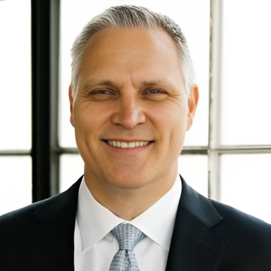 John Wilfong wearing a dark suit, white shirt, and a light gray tie, standing in front of large windows.