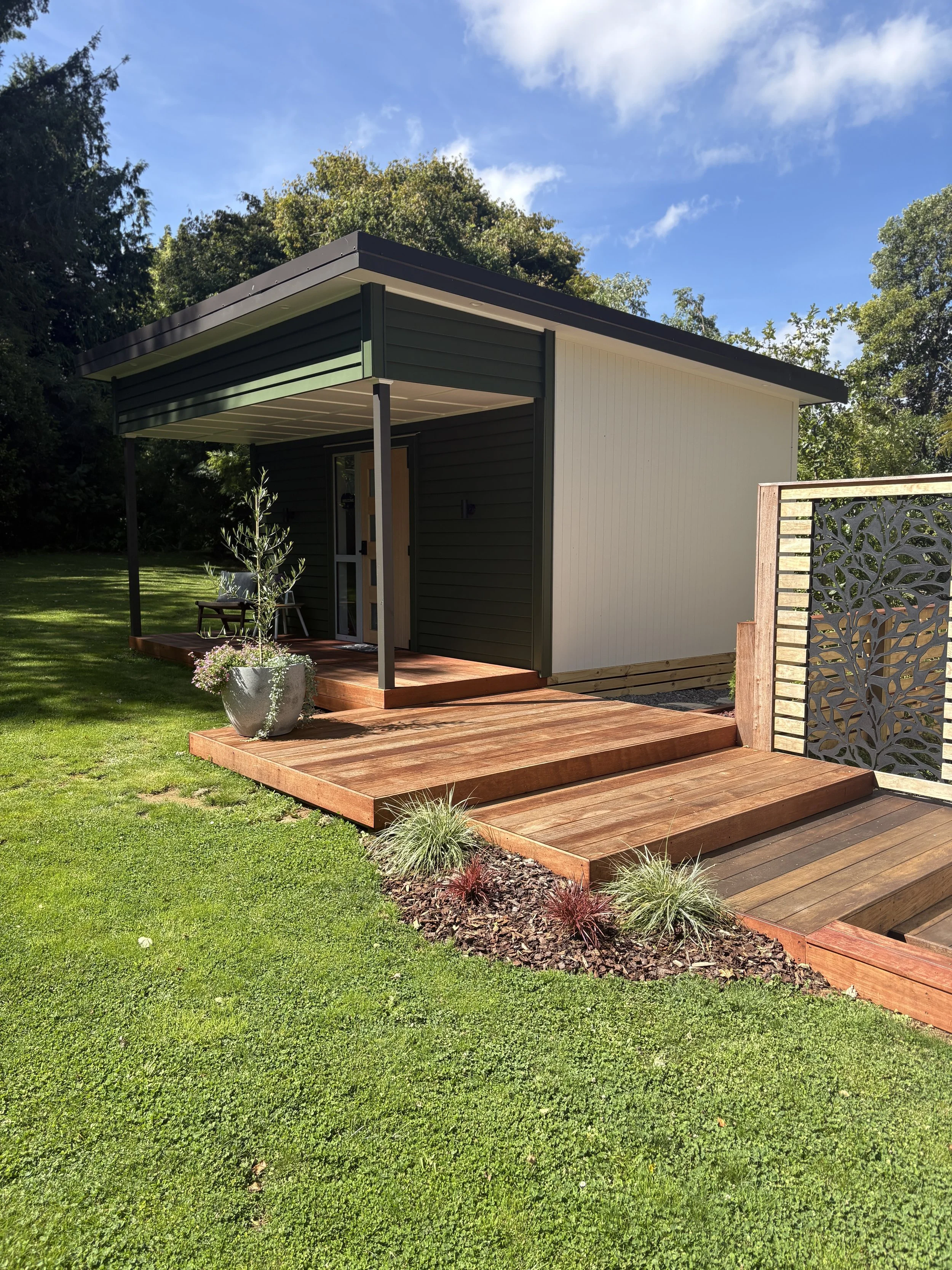 A small modern house with dark green and white exterior walls, a metal roof, a wooden porch with steps, and a lawn with a plant in a large pot, under a partly cloudy blue sky.